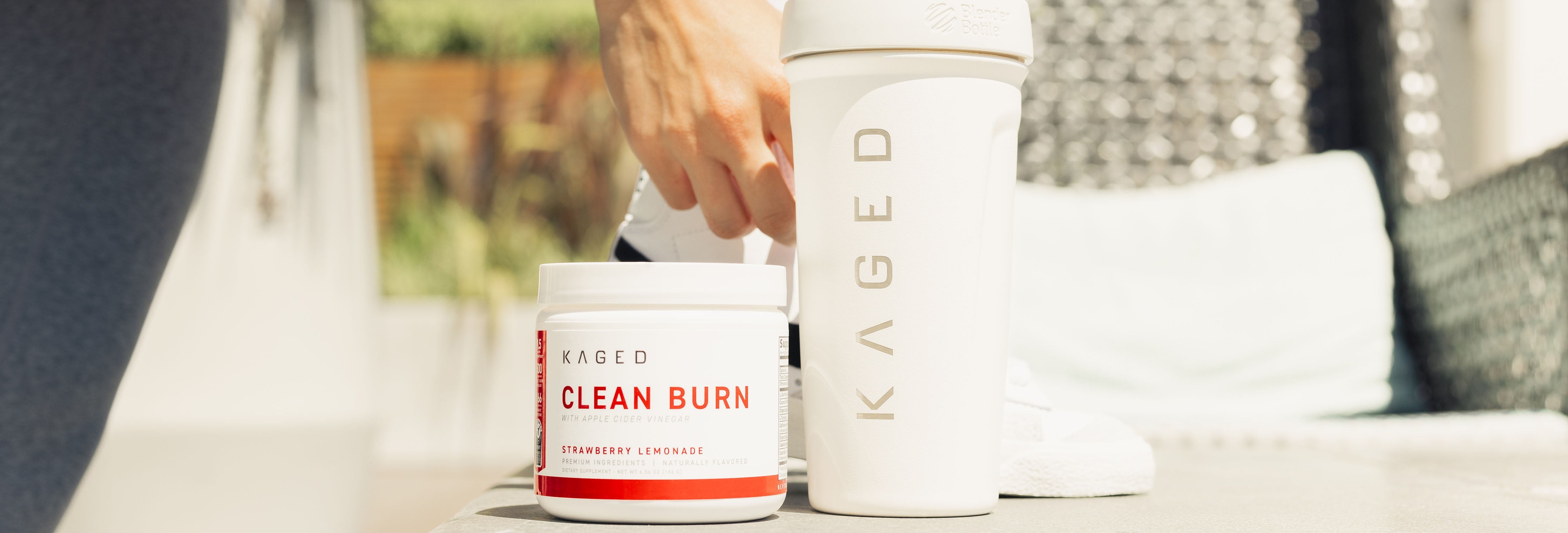 Front view of Kaged Clean Burn supplement jar next to white Kaged shaker on outdoor bench with hand reaching