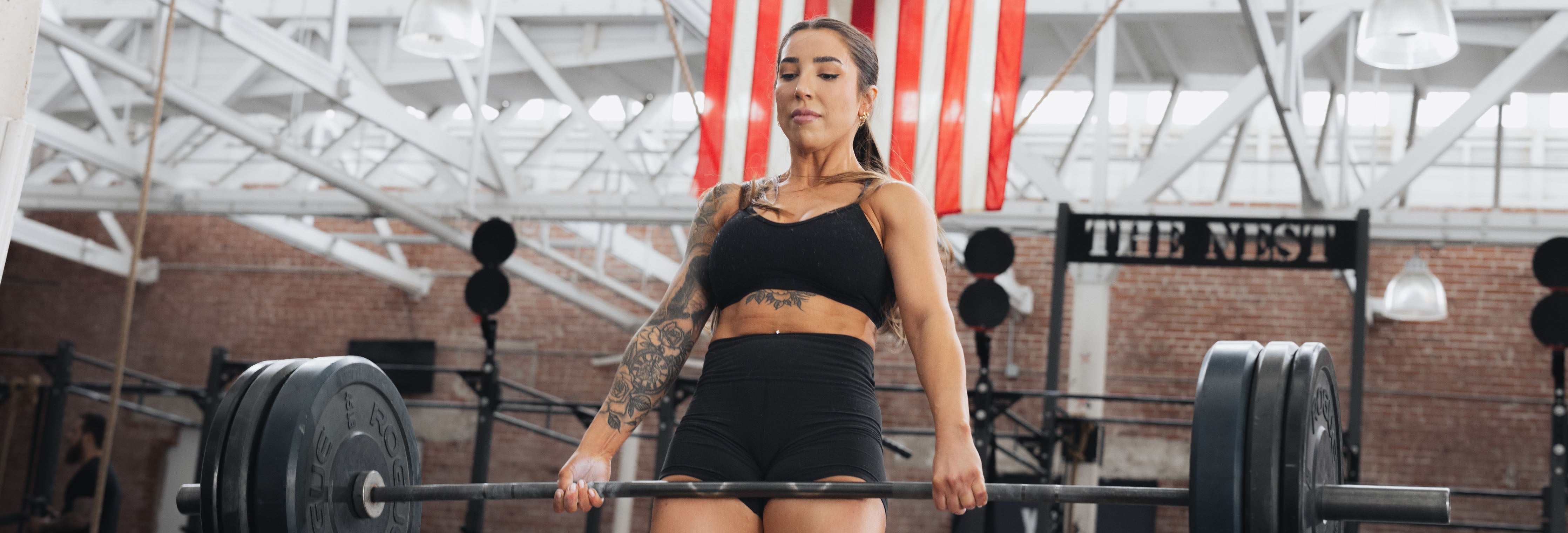 Female athlete deadlifting a heavily loaded barbell in an industrial-style gym with an American flag in the background