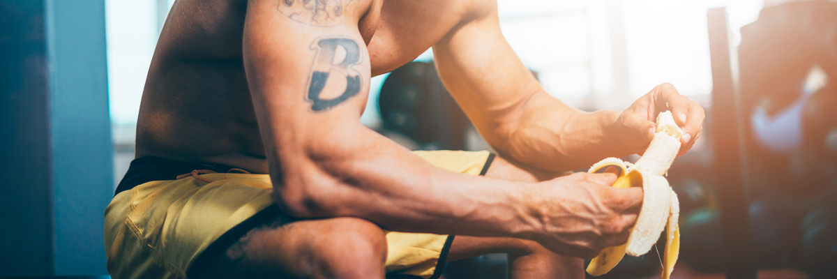 Athlete sitting in a gym peeling a banana between sets, shirtless with yellow shorts and a tattooed arm