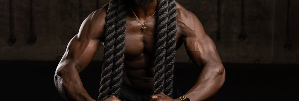 Shirtless muscular man gripping thick battle ropes in a dim gym setting