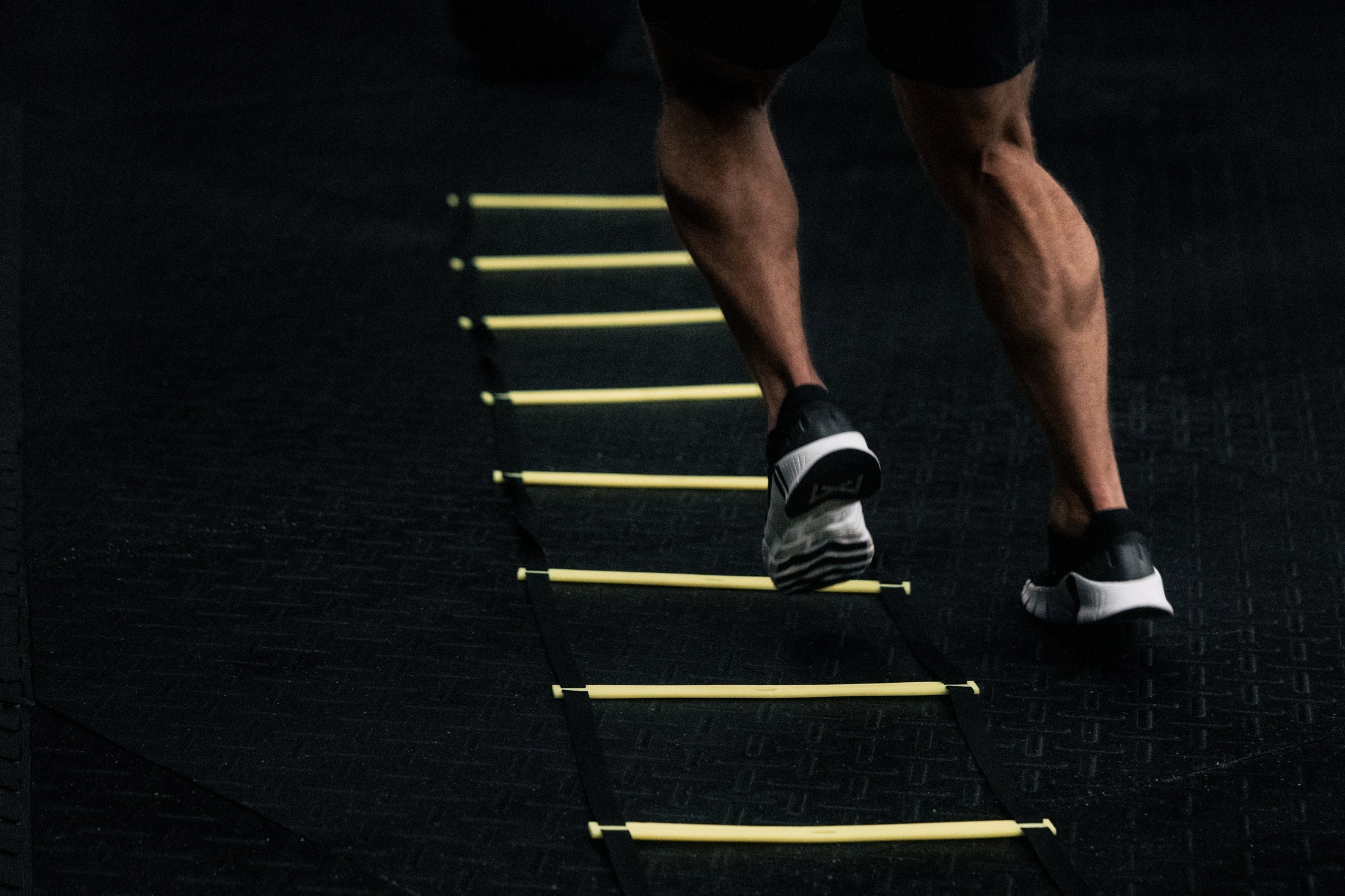 Person doing agility ladder drills on a black gym floor, highlighting defined calf muscles