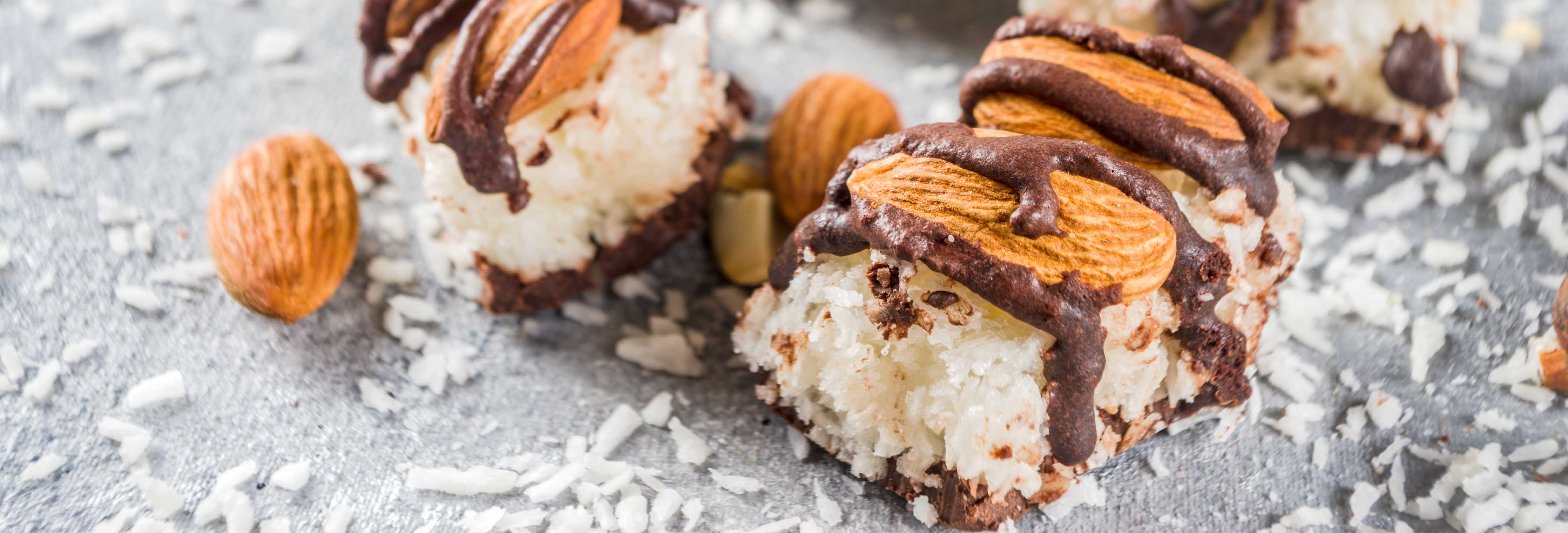Chocolate-drizzled coconut almond bites on a gray stone surface with scattered shredded coconut and whole almonds