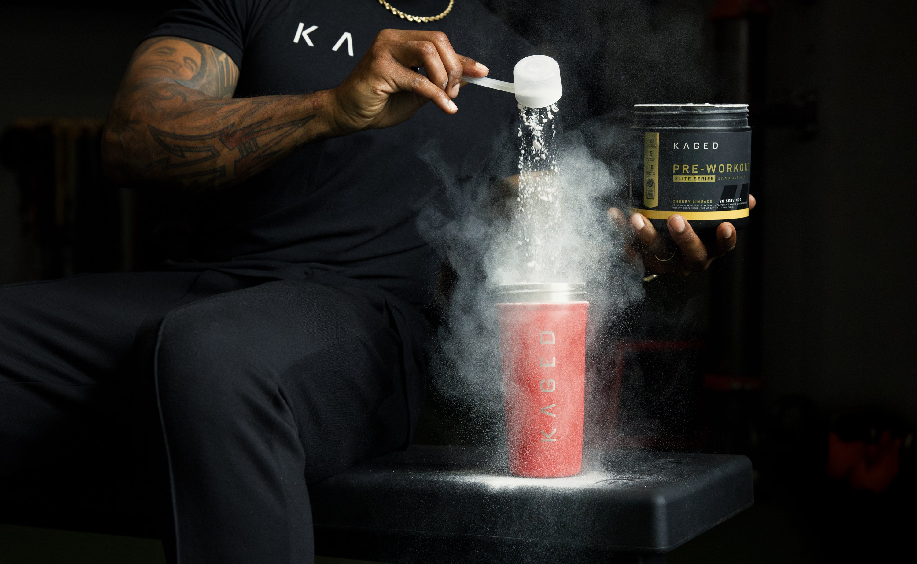 Athlete pouring a scoop of KAGED pre-workout powder from a tub into a red KAGED shaker, powder cloud rising in a dark gym setting