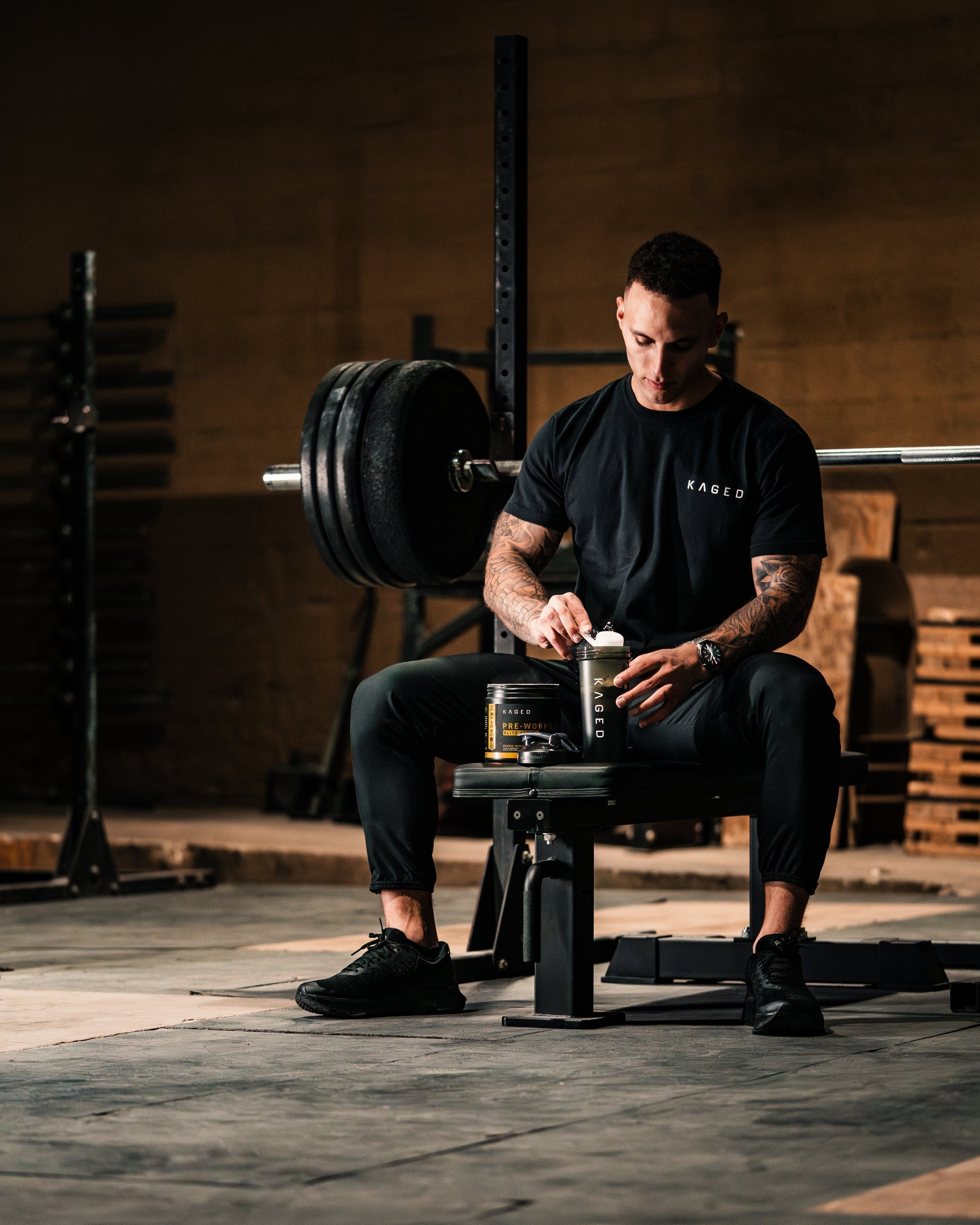 Man sitting on a gym bench preparing a KAGED pre-workout shaker with a tub of pre-workout and a loaded barbell behind him