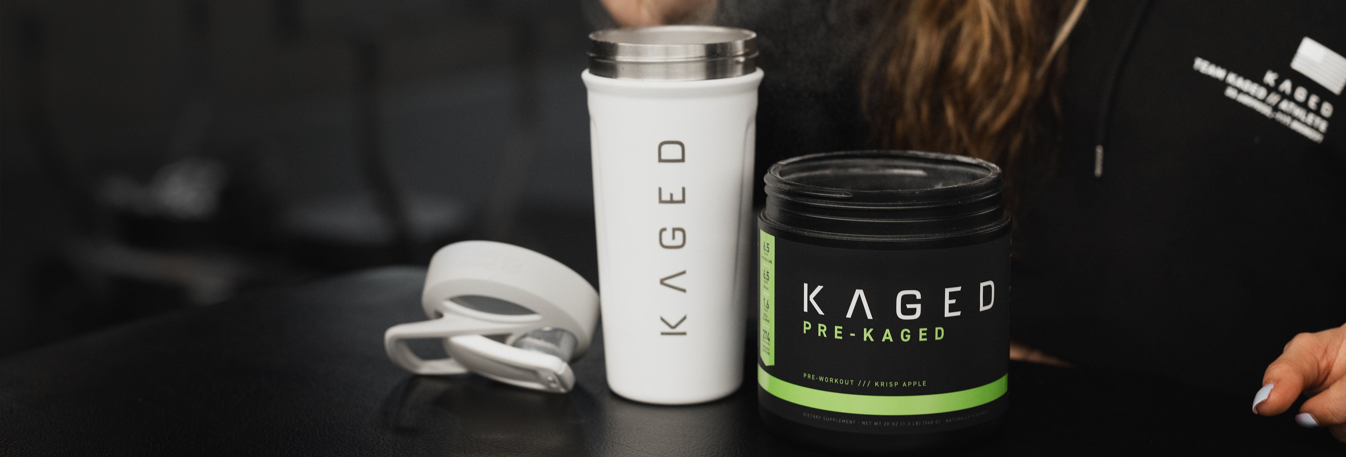 Front view of KAGED Pre-Kaged pre workout tub with green accent beside white KAGED shaker and lid on dark gym bench