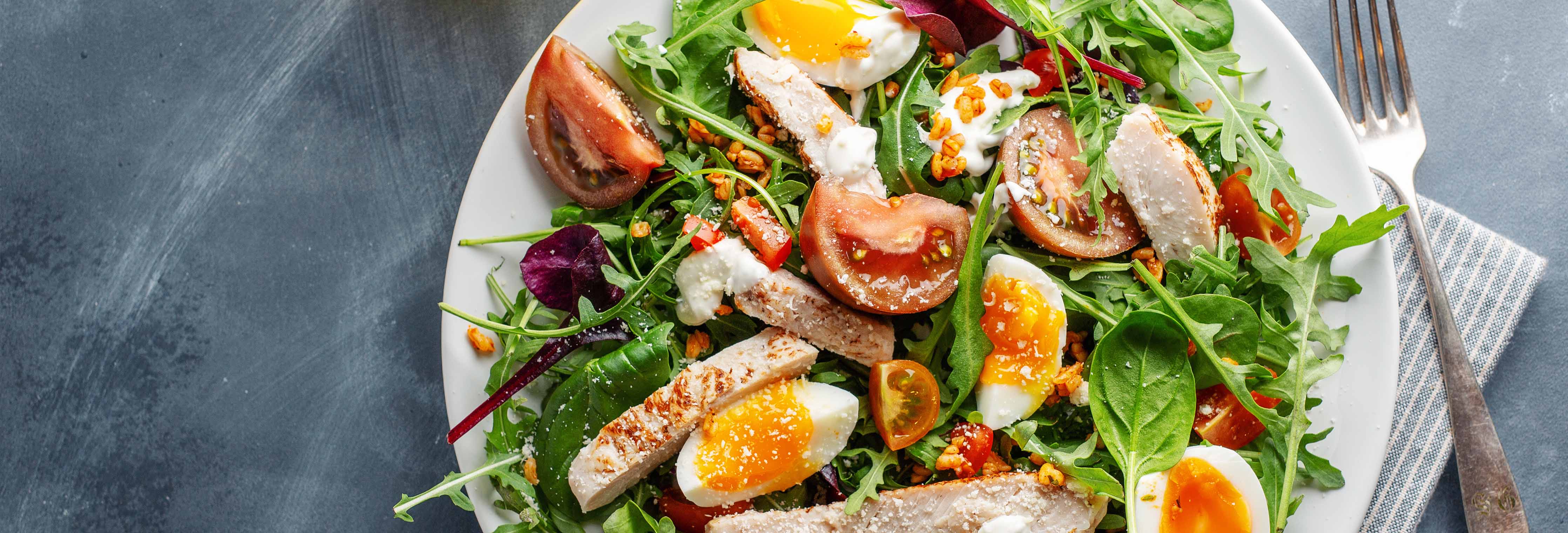 Grilled chicken and soft-boiled egg salad with arugula and cherry tomatoes on a white plate over a dark tabletop