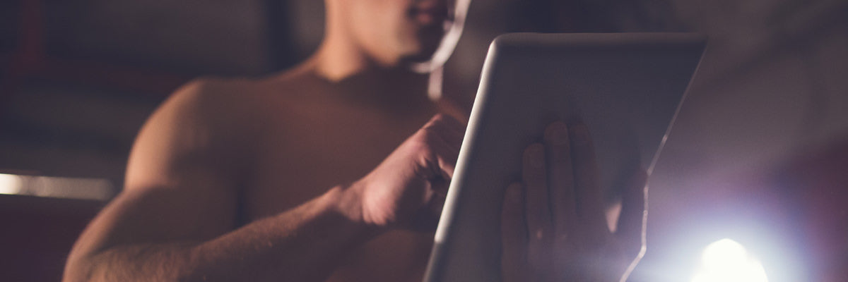 Shirtless man tapping a tablet in a dimly lit gym, focused on tracking his workout and sports nutrition