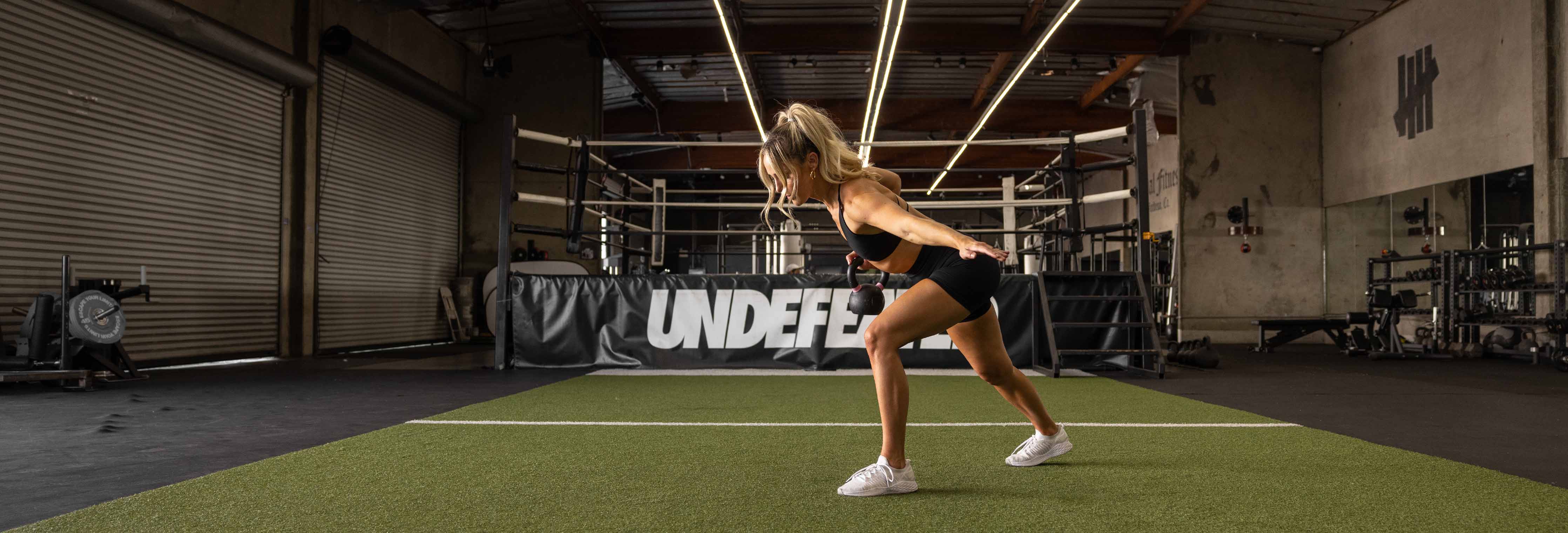 Woman doing a rear lunge on green turf in a gym, boxing ring behind her and large UNDEFEATED banner