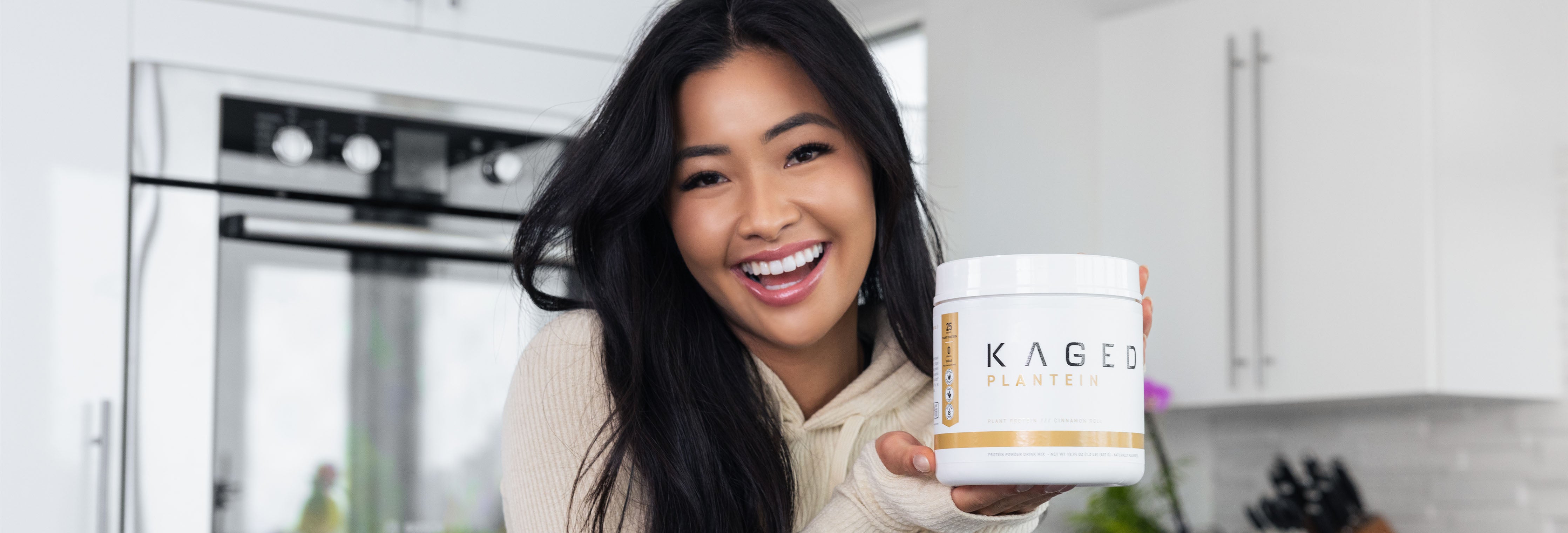 Front view of Kaged Plantein protein tub showcasing the label held by a smiling woman in a bright white kitchen