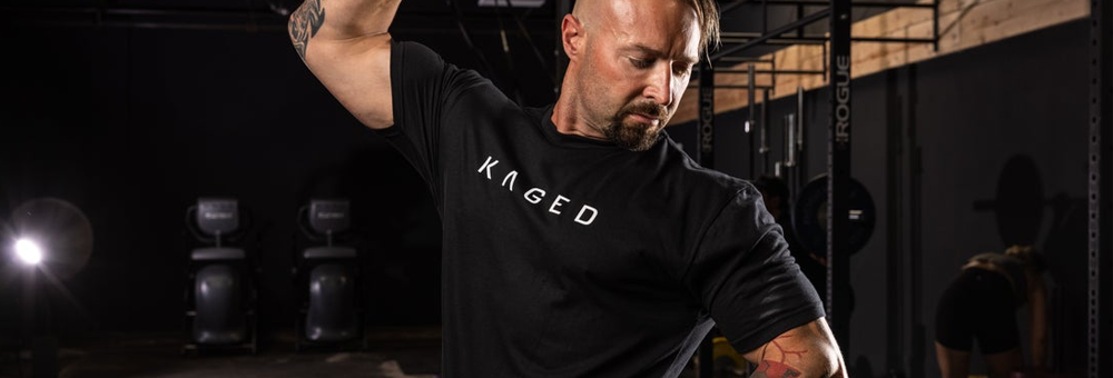 Athlete doing a dumbbell shoulder press in a dim gym wearing a black KAGED T-shirt