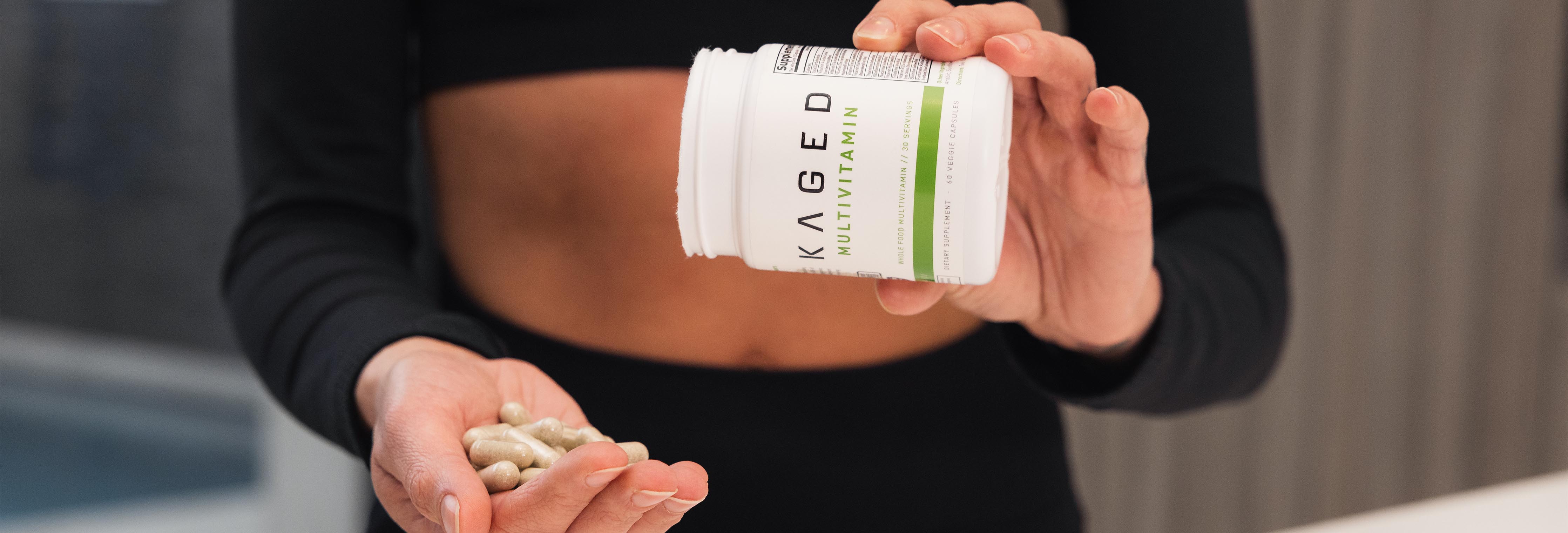 Woman in black workout top pouring KAGED Multivitamin capsules from bottle into her hand on a countertop