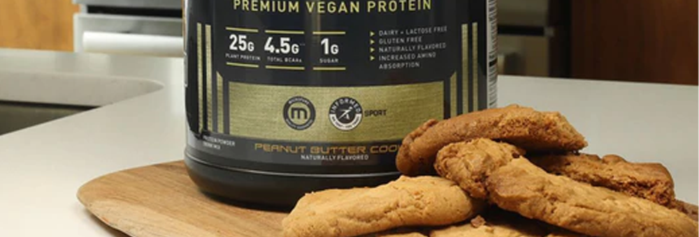 Front view of Kaged Premium Vegan Protein tub, Peanut Butter Cookie flavor, next to peanut butter cookies on a wooden board
