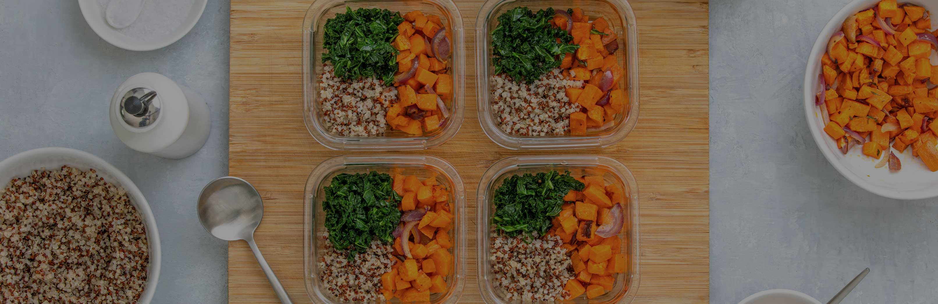 Four meal-prep containers with quinoa, roasted sweet potatoes, and sautéed greens on a wooden cutting board, top-down view