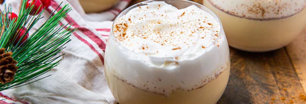 Two glasses of protein eggnog topped with whipped cream and grated nutmeg on a wooden surface with holiday pine sprig