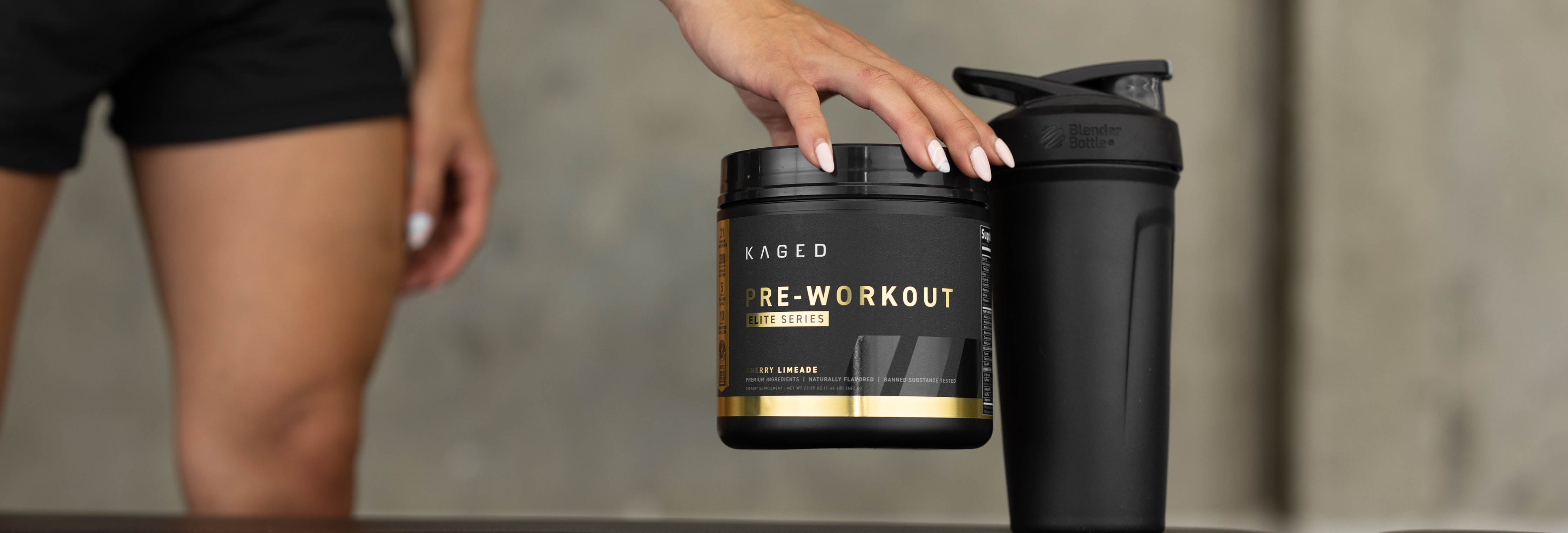 Front view of Kaged Pre-Workout tub held by hand beside a black shaker bottle on gym surface with blurred legs in background