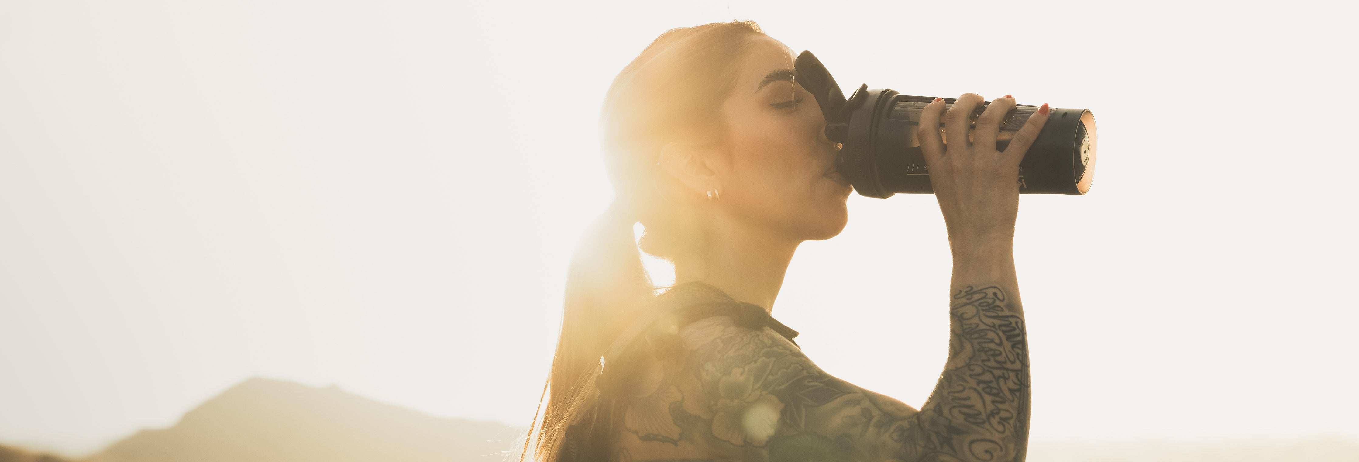 Woman drinking from a supplement shaker bottle at sunrise outdoors with sun flare
