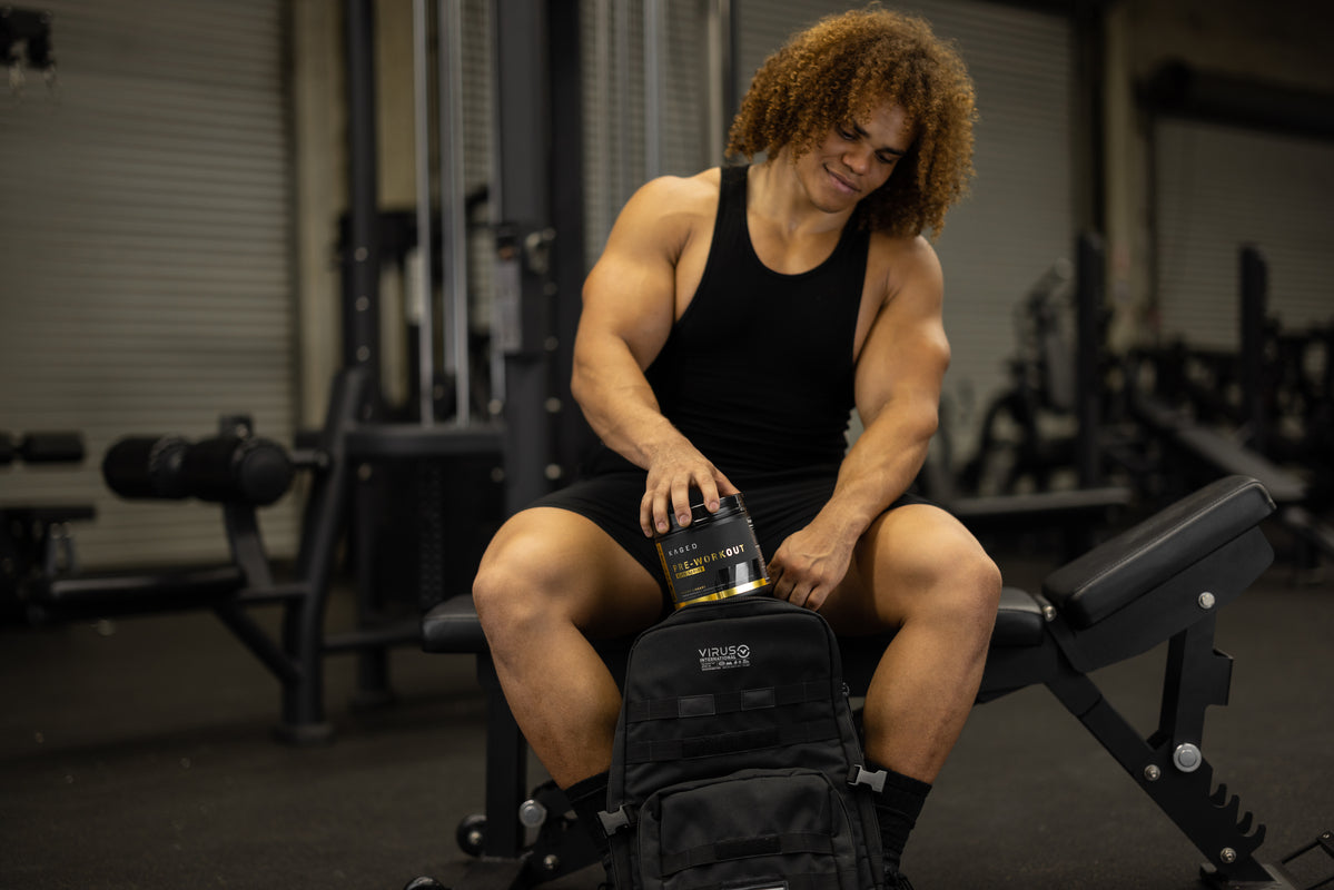 Athlete placing a KAGED pre workout tub into a black gym backpack while seated on a bench in a weight room