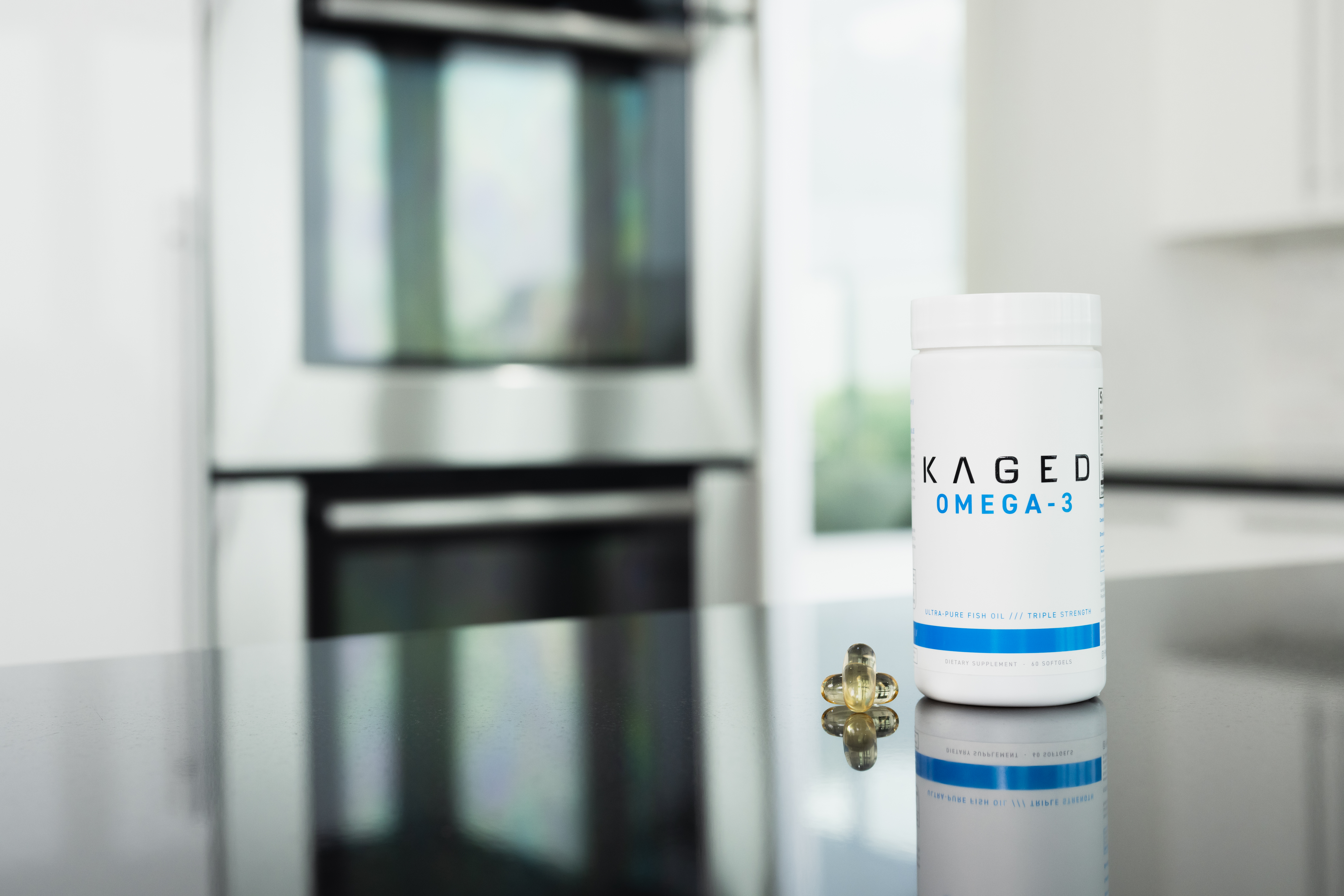 Front view of KAGED Omega-3 supplement bottle with three softgel capsules on a reflective kitchen countertop