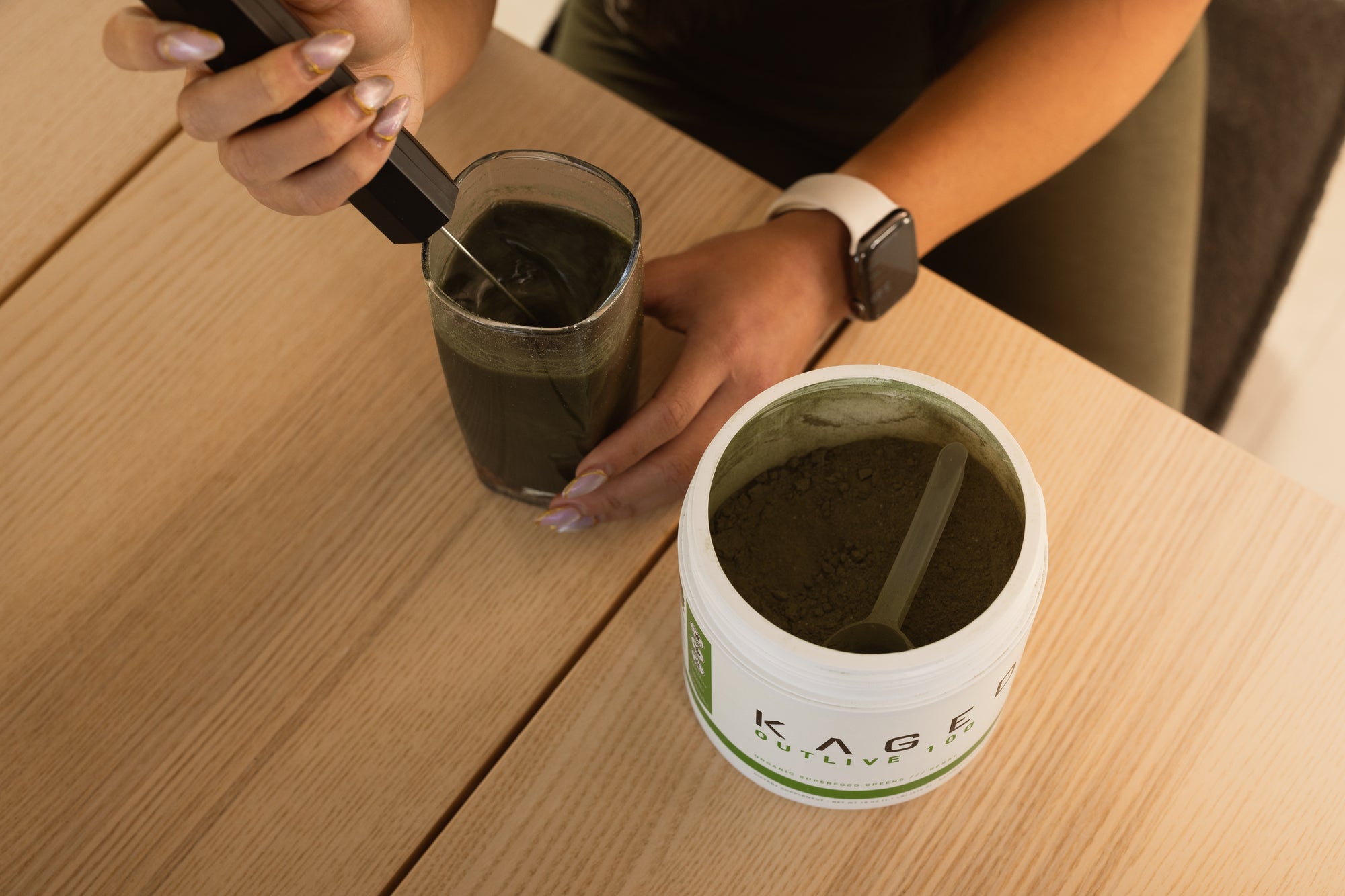 Person mixing a green Organic Greens drink with a frother beside an open KAGED Greens tub on a wooden table