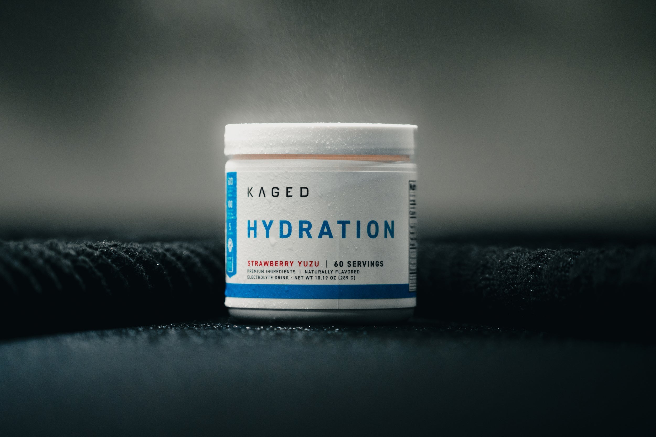 Front view of Kaged Hydration tub showing the front label reading Hydration, Strawberry Yuzu flavor and 60 servings on a dark textured blurred background