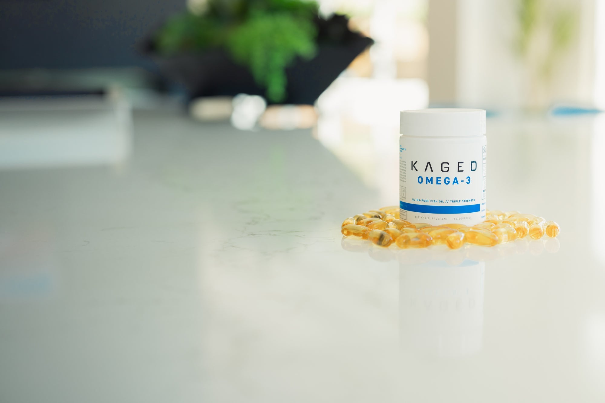 Front view of KAGED Omega-3 supplement jar with softgel capsules on a glossy white countertop and blurred background