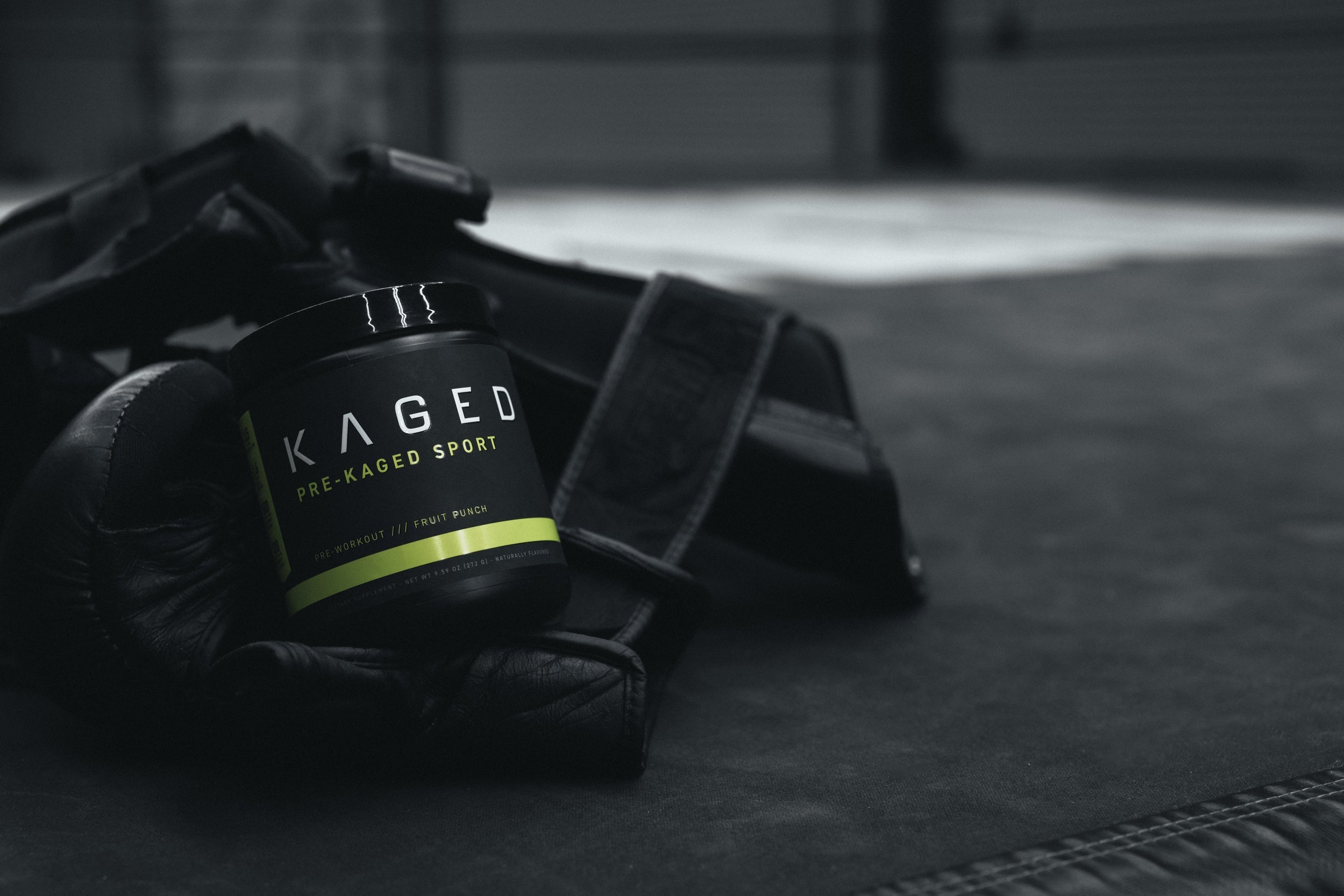 Angled front view of KAGED Pre-Kaged Sport pre-workout tub showing the label and Fruit Punch flavor on boxing gloves and a gym mat
