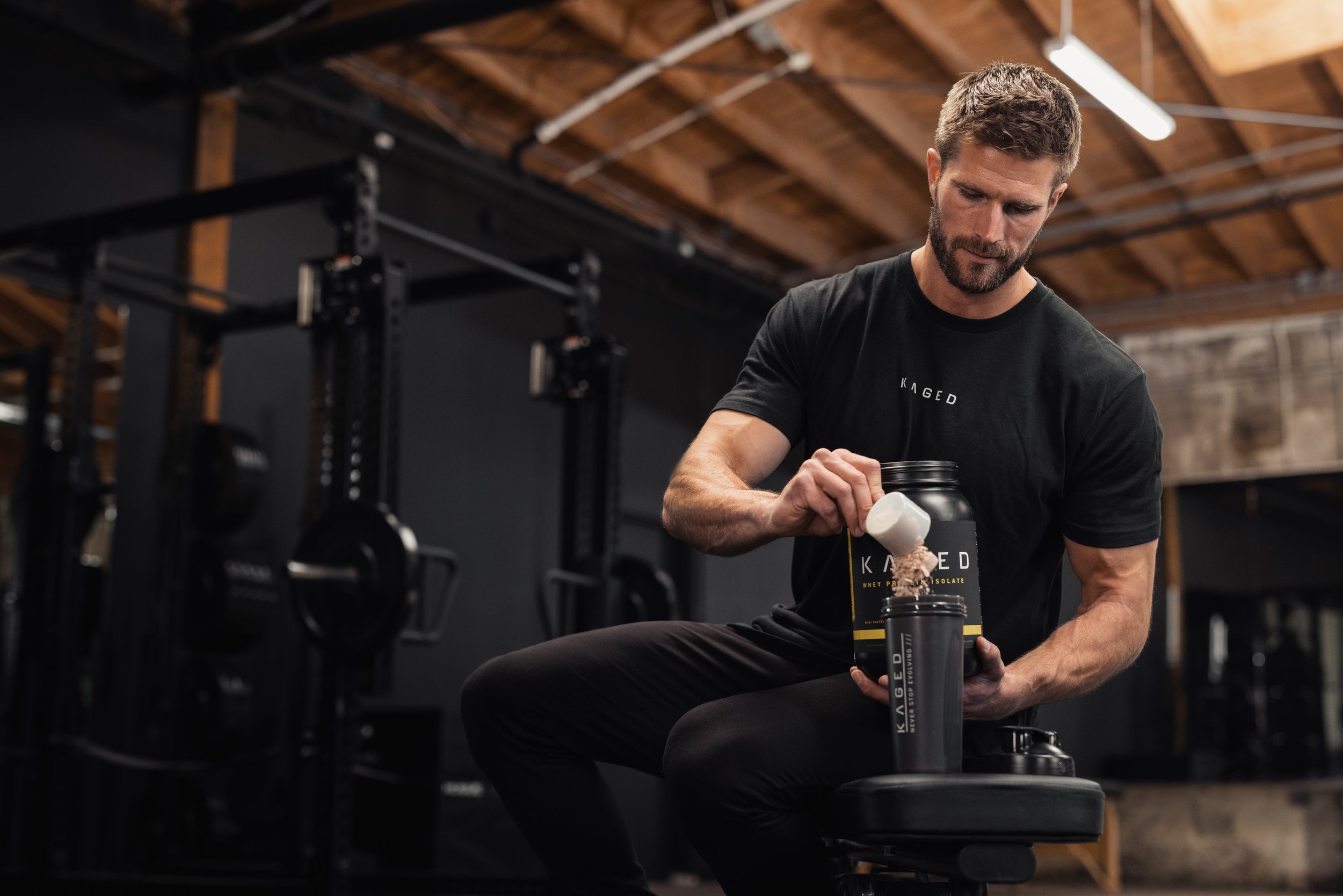 Man in a gym pouring KAGED whey protein powder from a tub into a black KAGED shaker while seated on a bench with a weight rack behind him