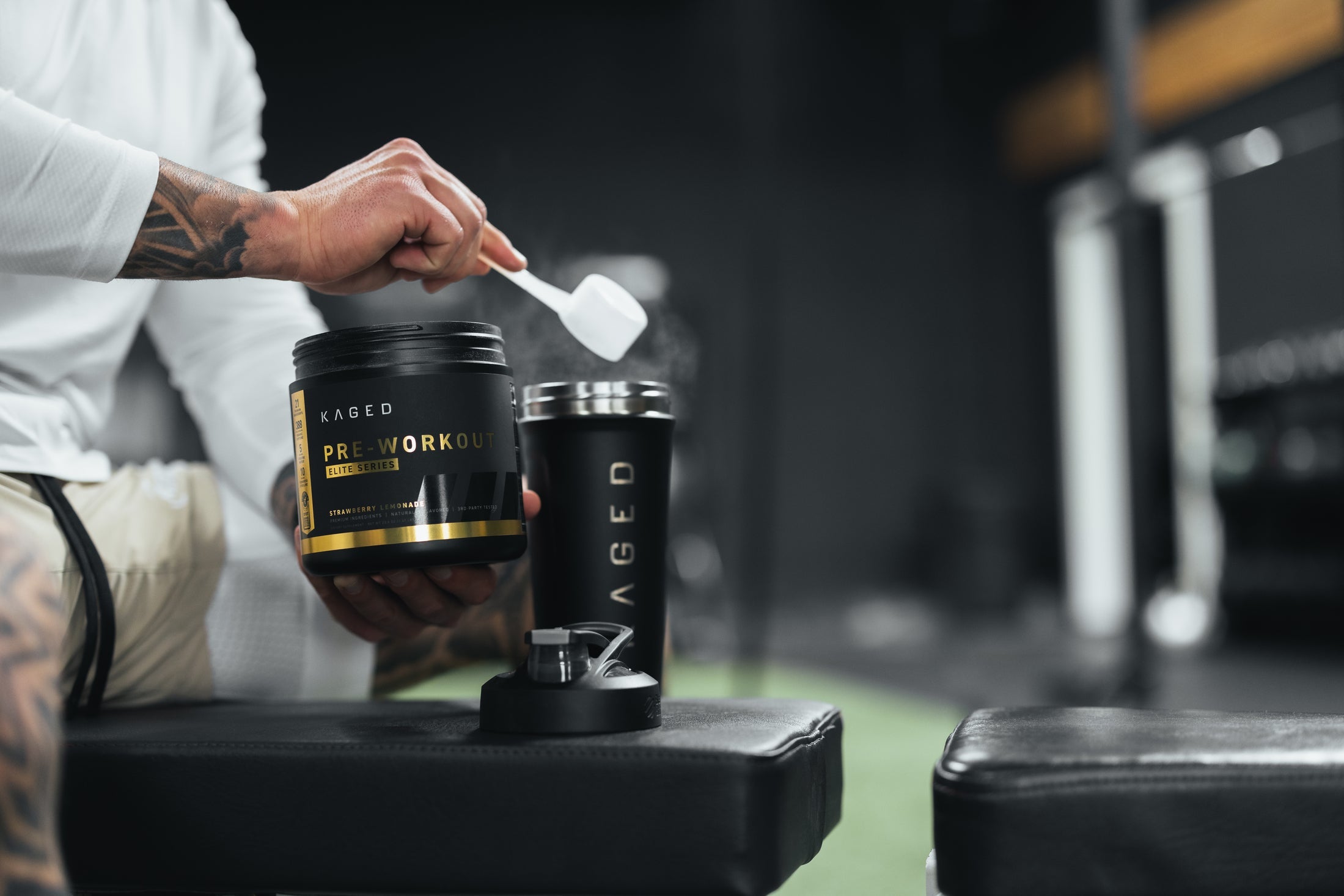 Close-up of KAGED Pre-Workout tub showcasing a scoop of powder and the product label with a KAGED shaker and blurred gym bench background
