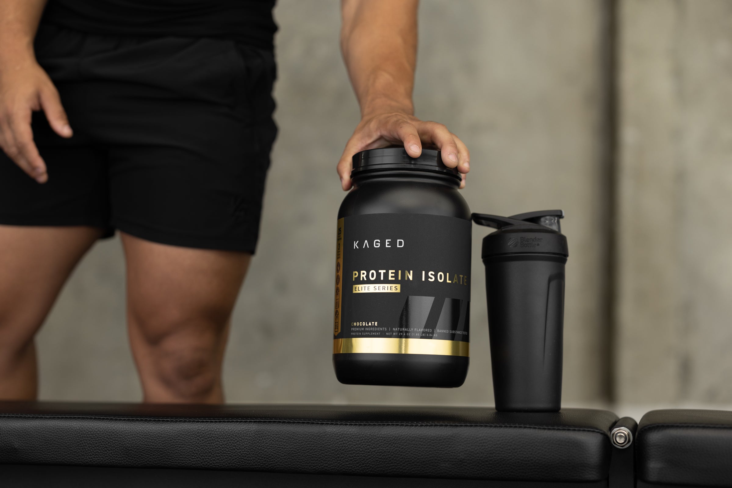 Front view of Kaged Protein Isolate tub showcasing black and gold packaging with shaker on a gym bench and athlete legs