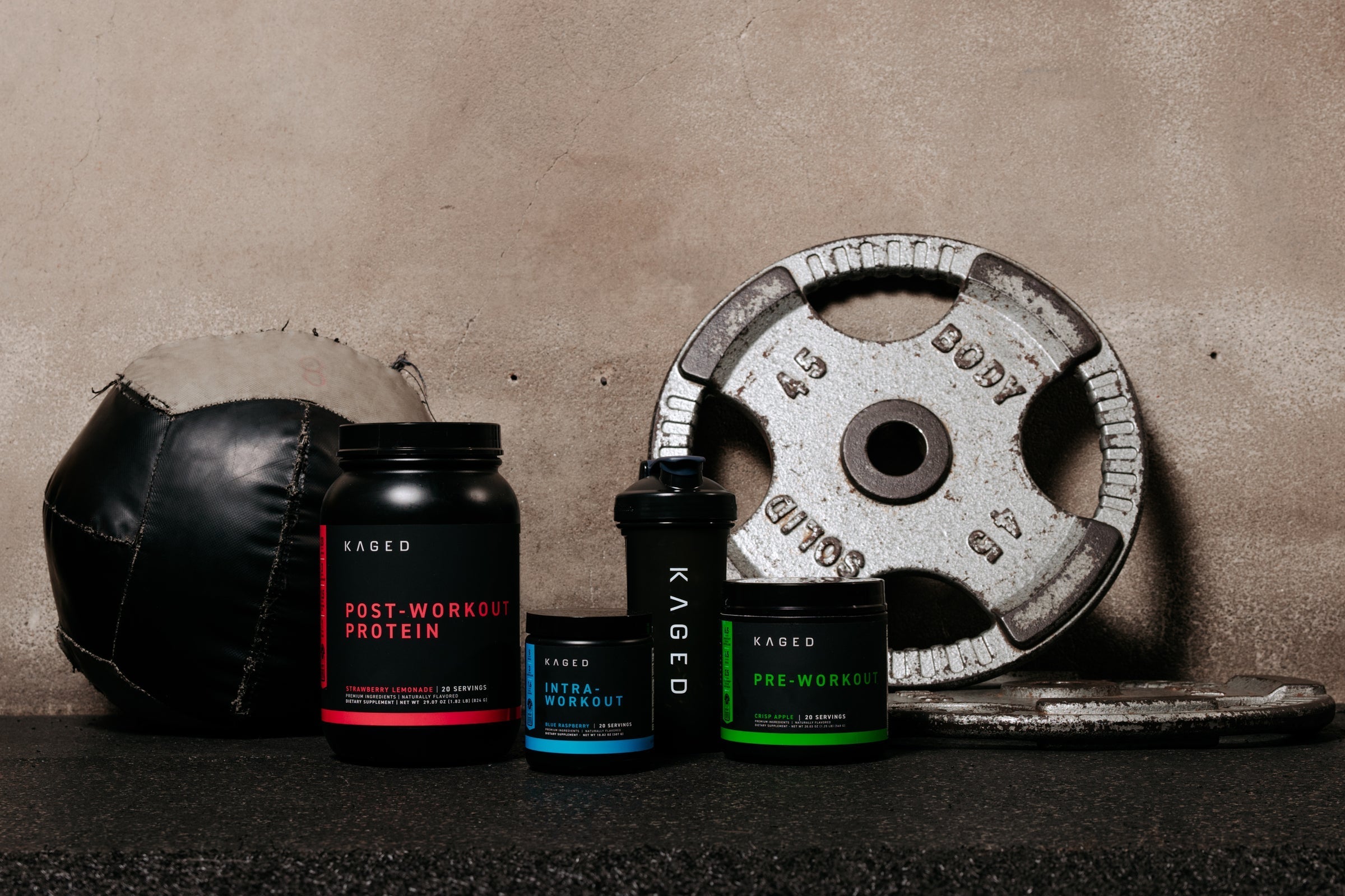 Front view of KAGED PIP stack: pre-, intra-, and post-workout tubs with shaker on gym floor and 45 lb weight plate