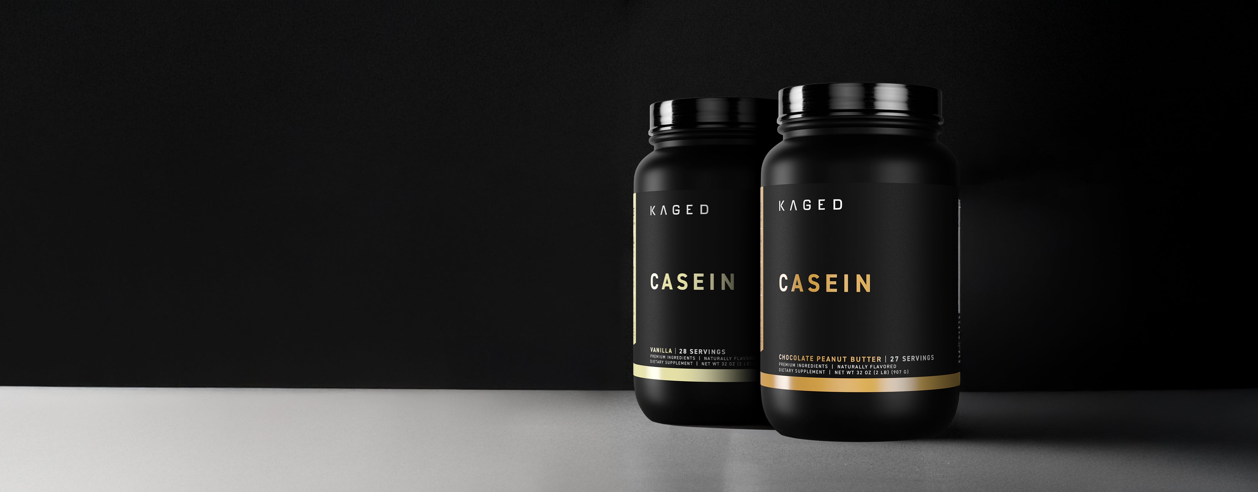 Front and side view of Kaged Casein protein tubs, matte black jars with gold CASEIN label on gray surface and dark background