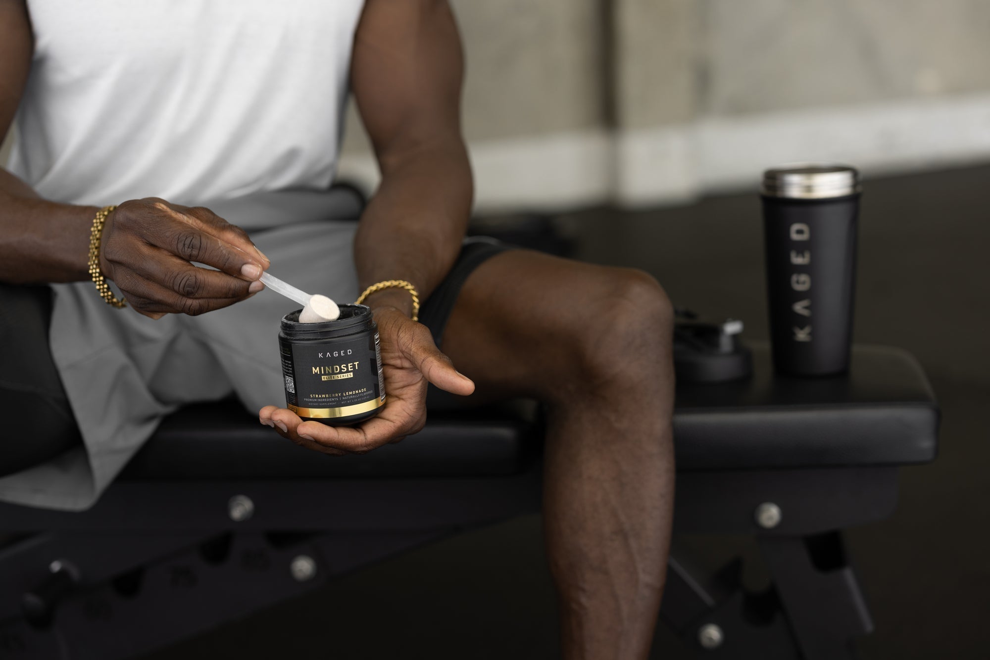 Person scooping KAGED Mindset Elite powder from a jar while seated on a gym bench with a KAGED shaker cup nearby
