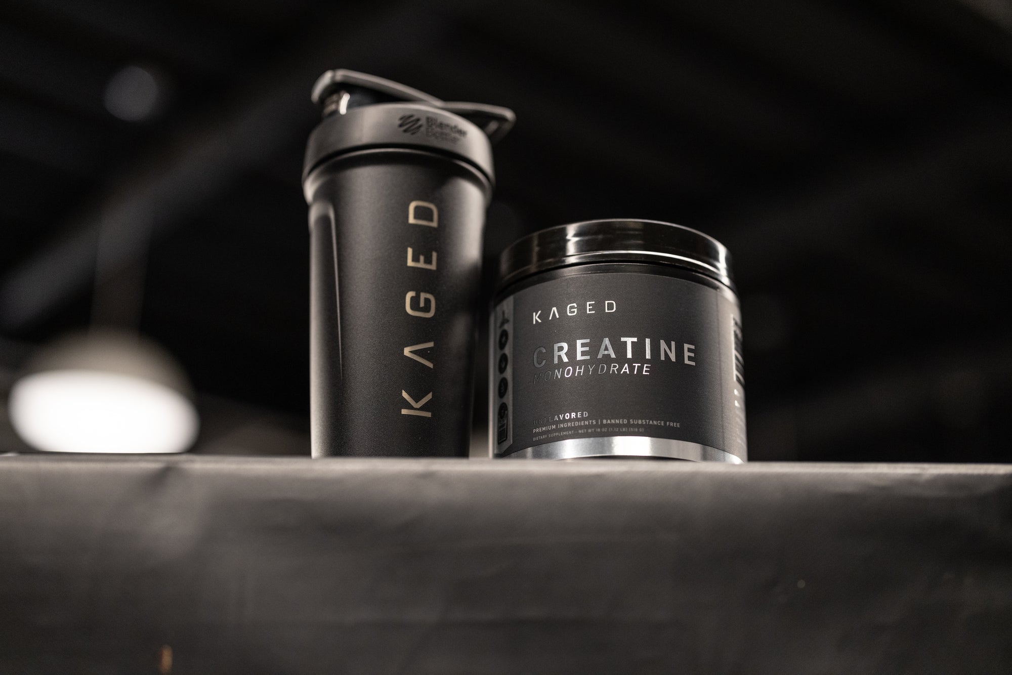 Low-angle of KAGED Creatine Monohydrate tub and black shaker showcasing label and gold logo with blurred gym background
