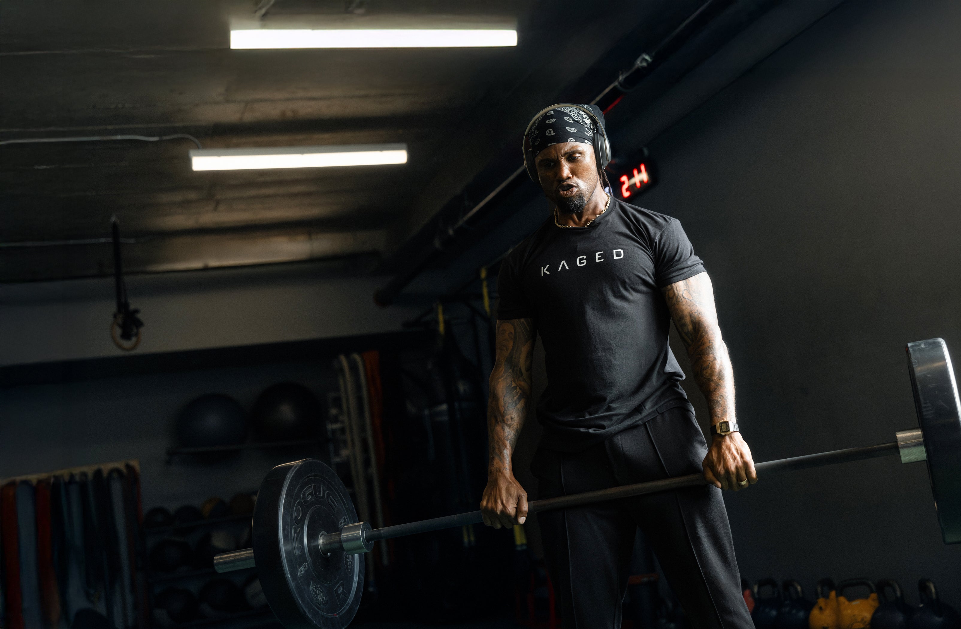 Man performing a deadlift in a dim gym wearing a black KAGED T-shirt, bandana, and headphones