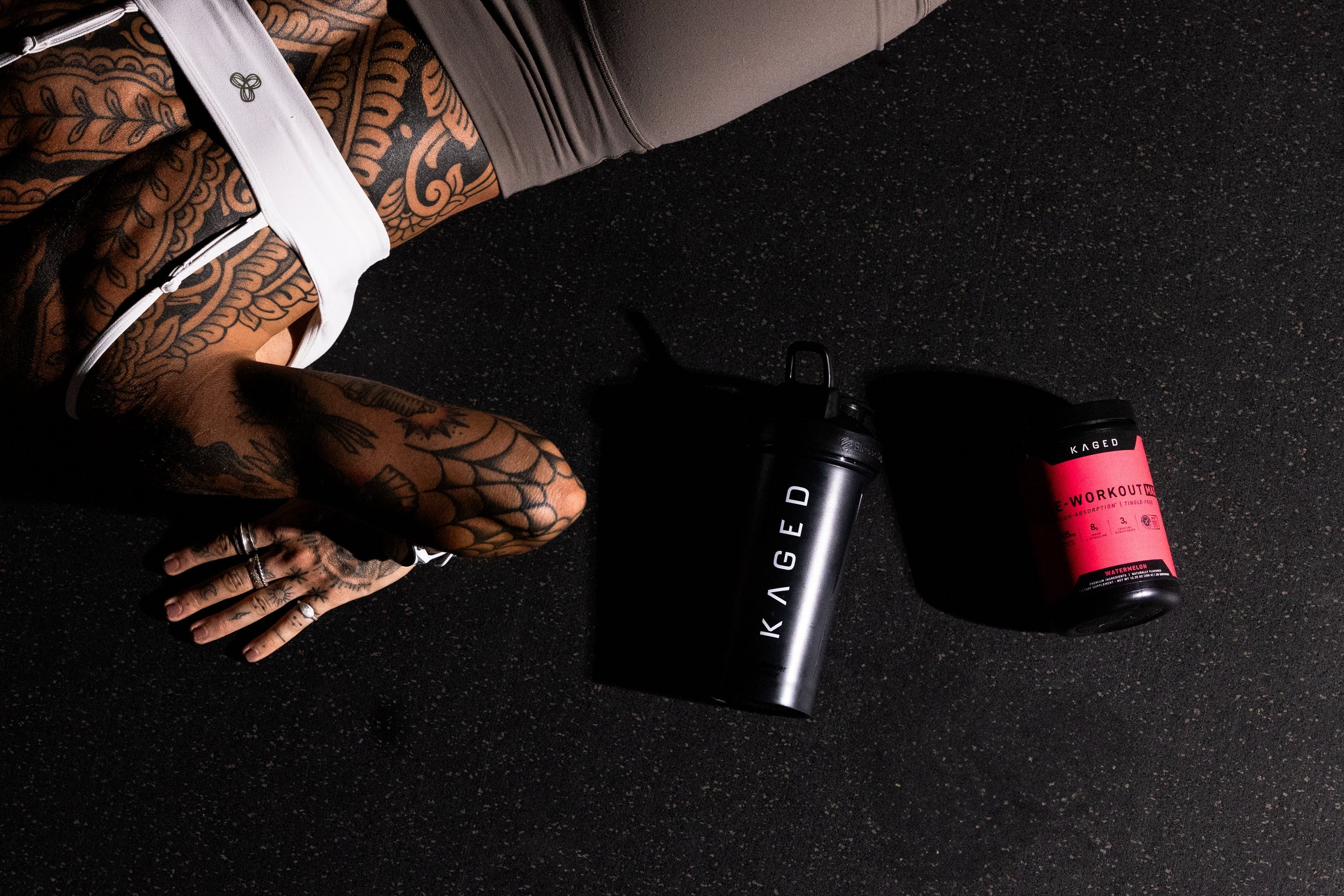 Tattooed person resting on a gym floor next to a black KAGED shaker and a pink-labeled Pre Workout Max tub