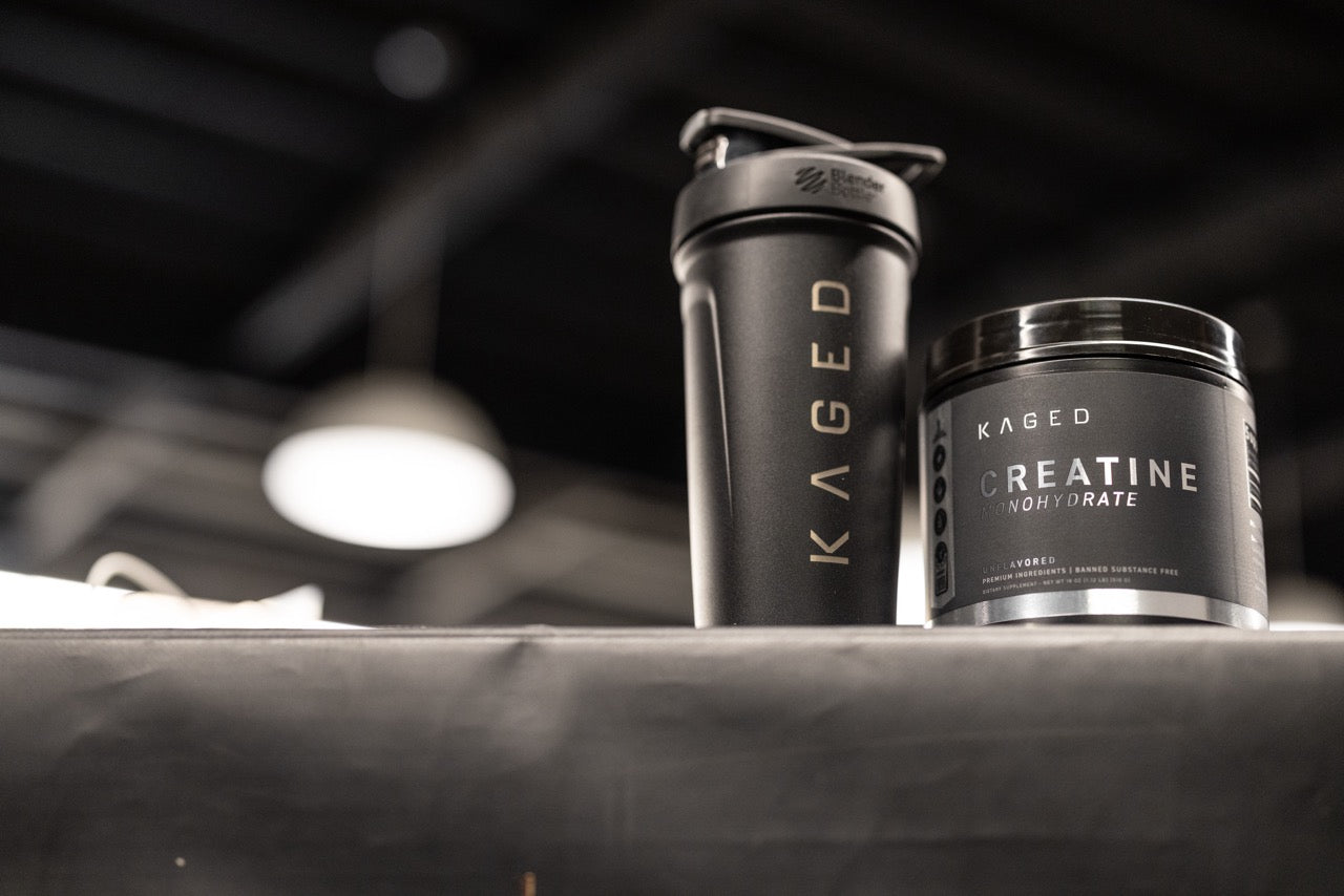 Low-angle view of KAGED creatine monohydrate tub and KAGED shaker showcasing black label and logo with blurred gym ceiling background