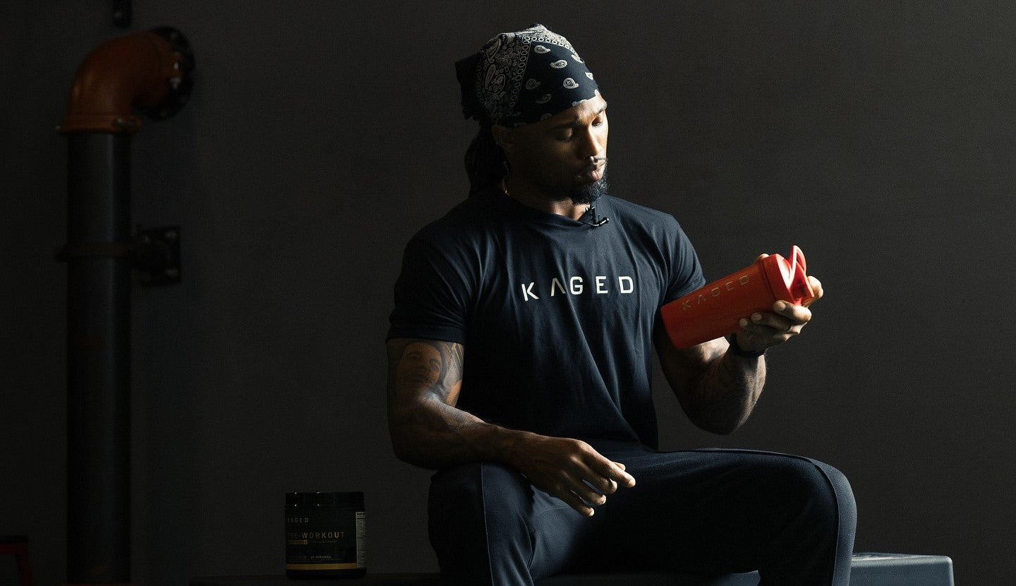 Man in black KAGED t-shirt holding and inspecting a red shaker bottle while seated in a dim gym with a pre-workout tub on the floor