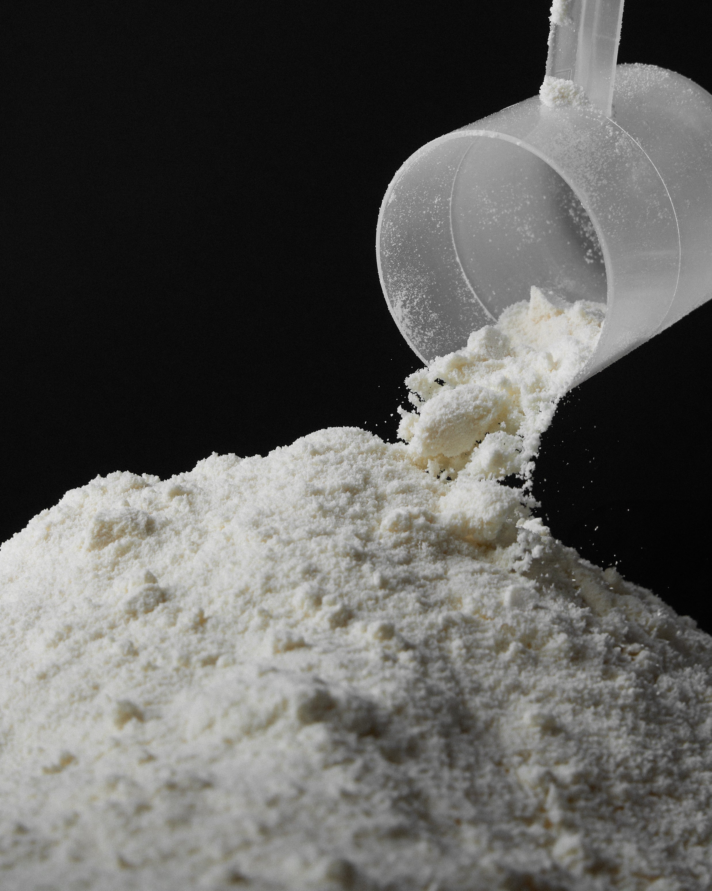 Close-up of a clear plastic scoop pouring white protein powder onto a pile on a black background