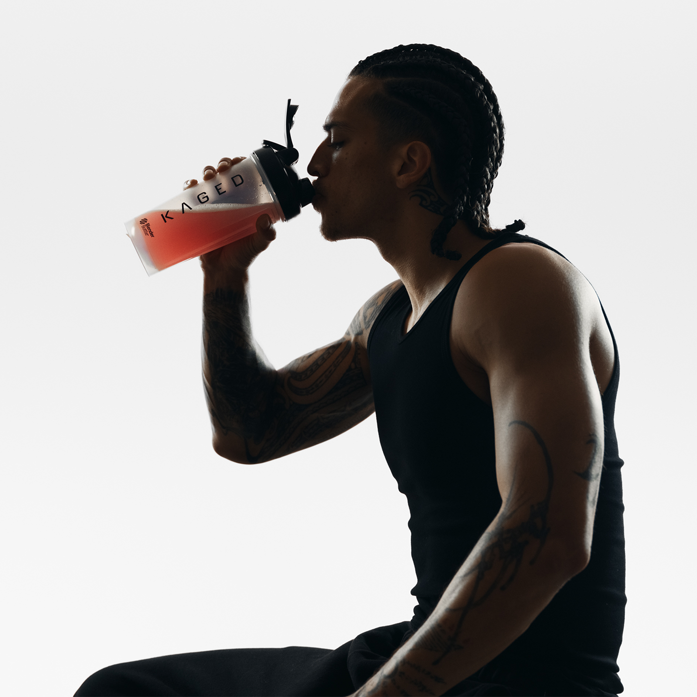 Man in black tank top drinking pink supplement from a KAGED shaker bottle against a white background