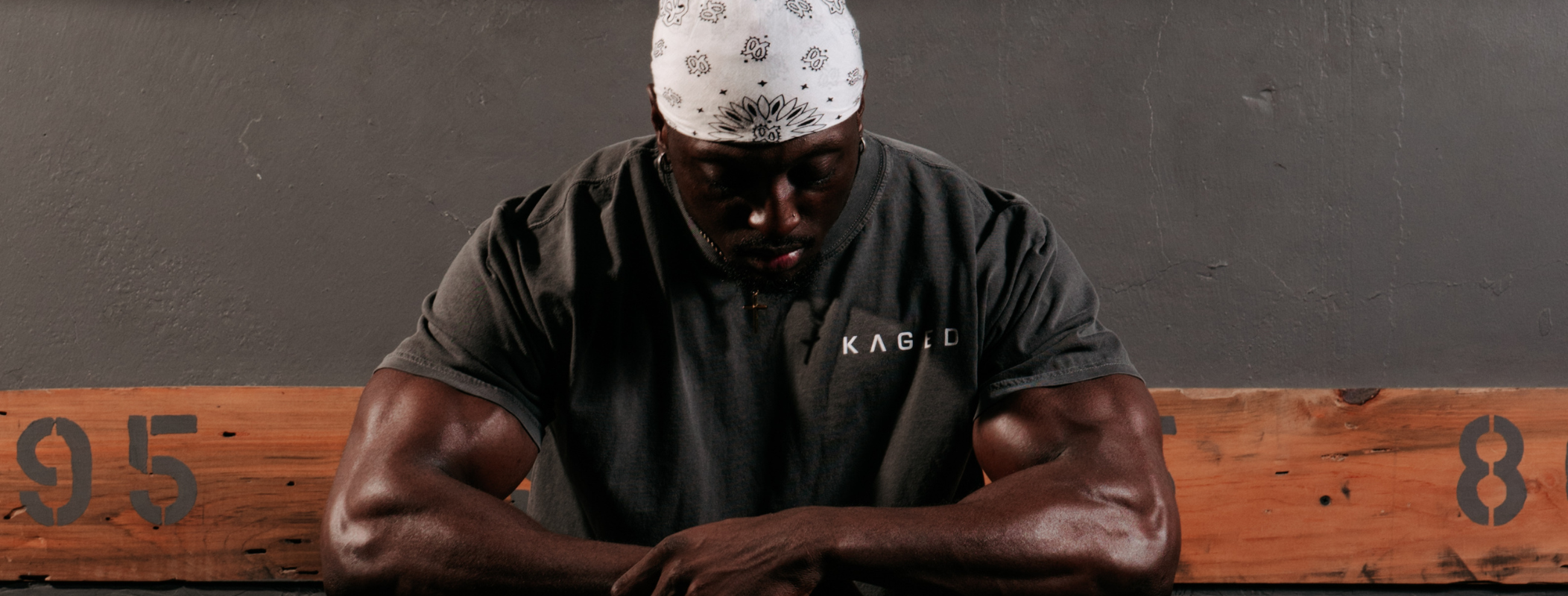 Muscular man wearing a KAGED t-shirt and bandana resting on a wooden bench against a gray wall, head bowed