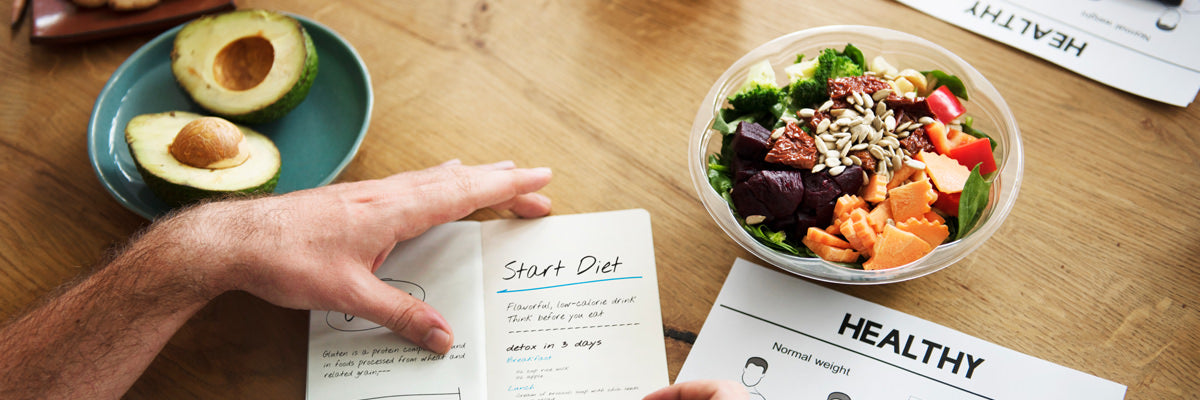 Person writing Start Diet notes beside avocado halves and a salad bowl on a table with a sheet titled Healthy