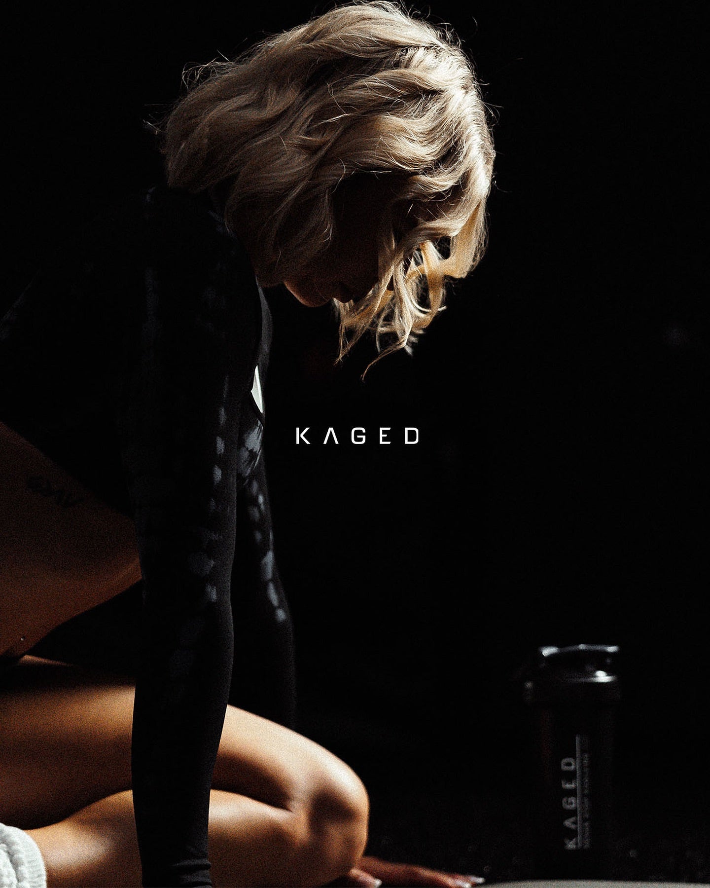 Woman resting post-workout with Kaged shaker bottle by her side in a dimly lit gym.