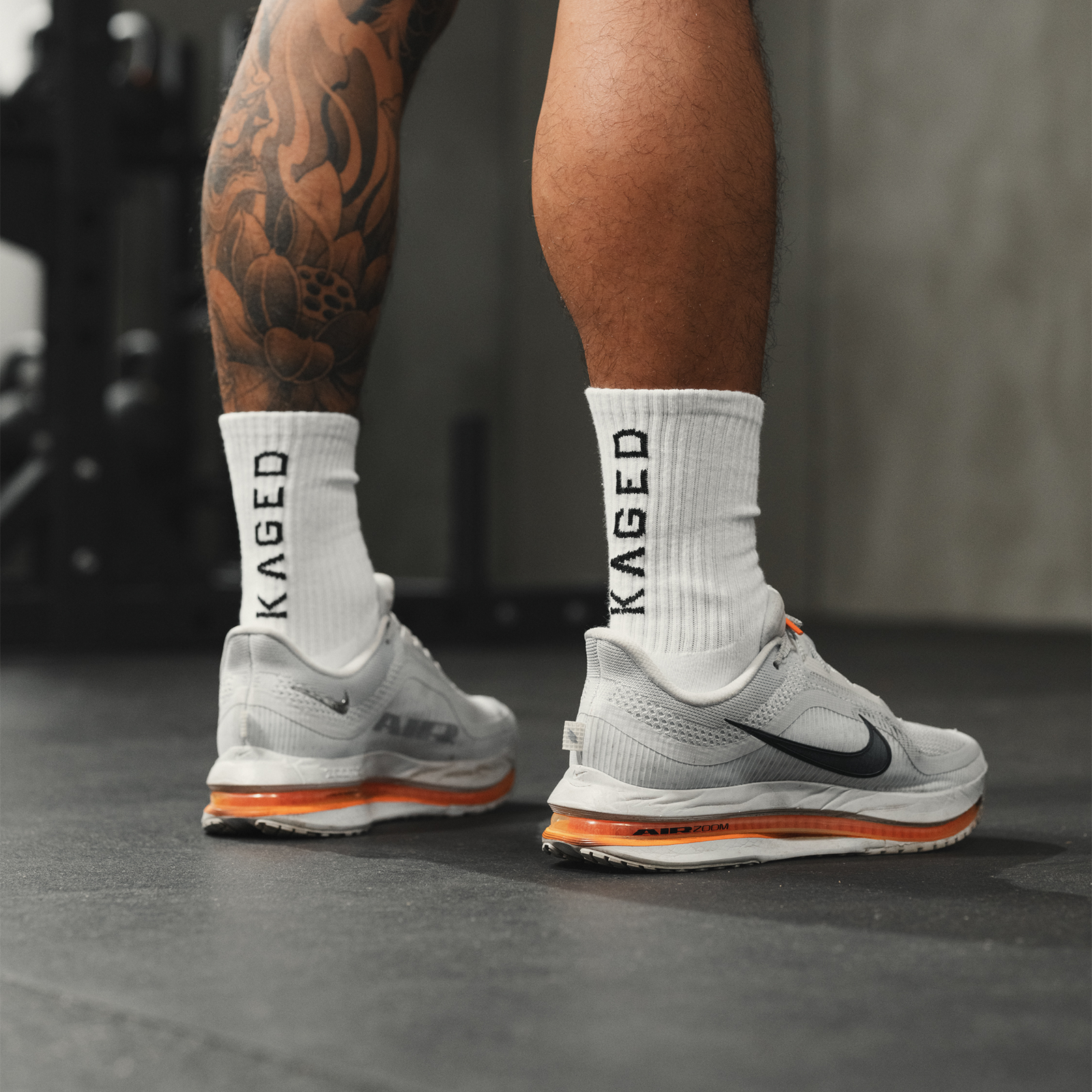 Rear view of white Ascent Performance socks on model showcasing vertical KAGED logo and ribbed crew height in gym setting