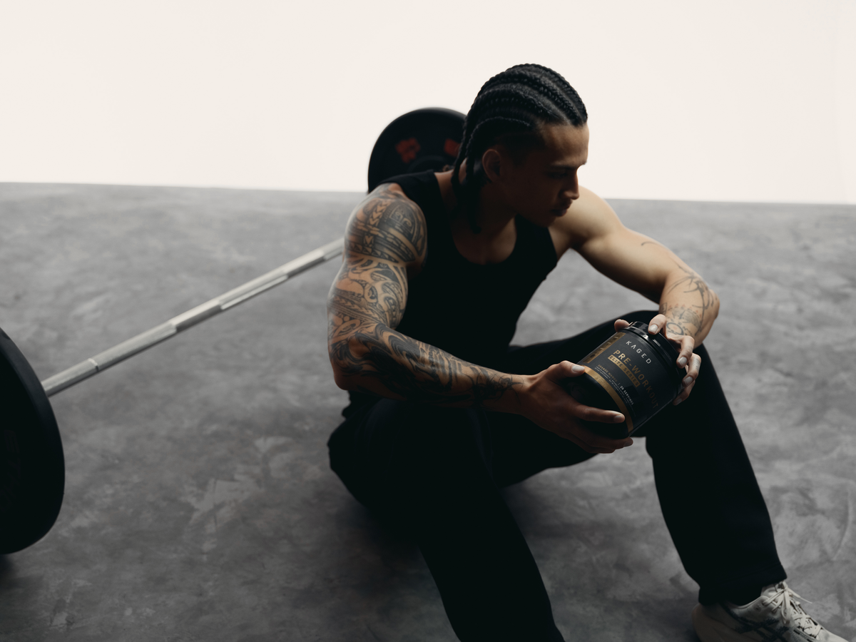 Athlete sitting on a gym floor holding a KAGED Pre-Workout tub beside a loaded barbell
