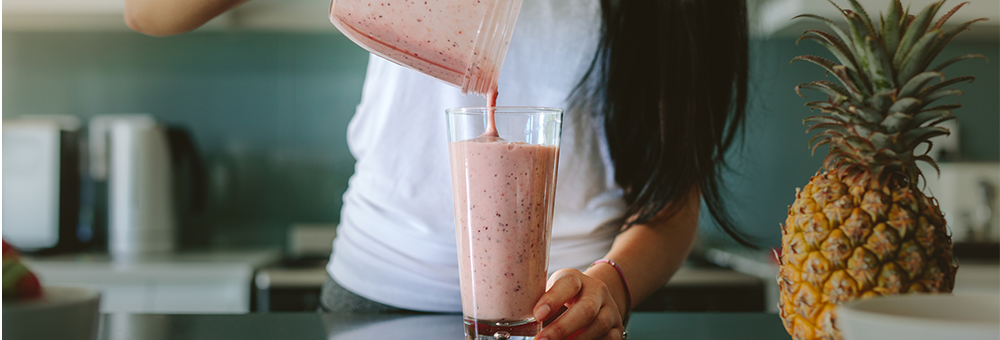 Person pouring a pink fruit smoothie into a tall glass on a kitchen counter with a pineapple nearby