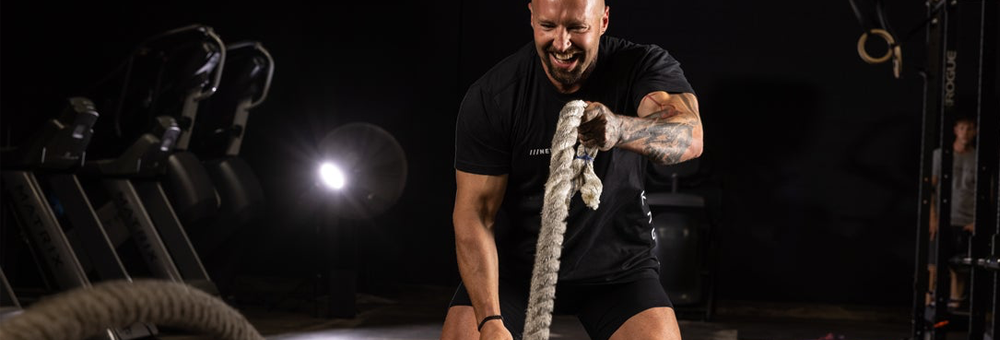 Trainer performing battle rope workout in a dimly lit gym with weight equipment in the background, smiling