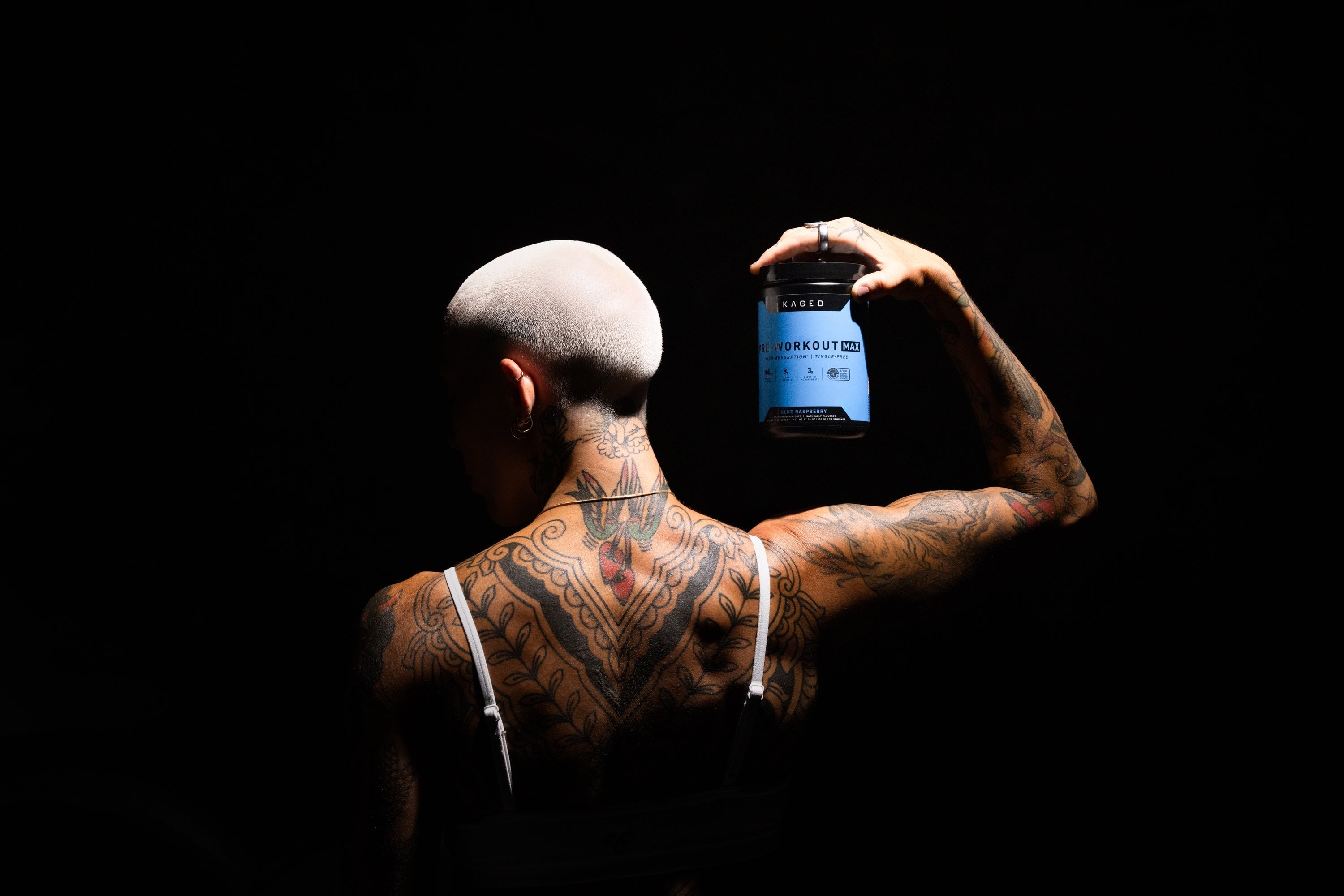 Rear view of KAGED Workout Max pre-workout tub held above a tattooed person's shoulder against a black background