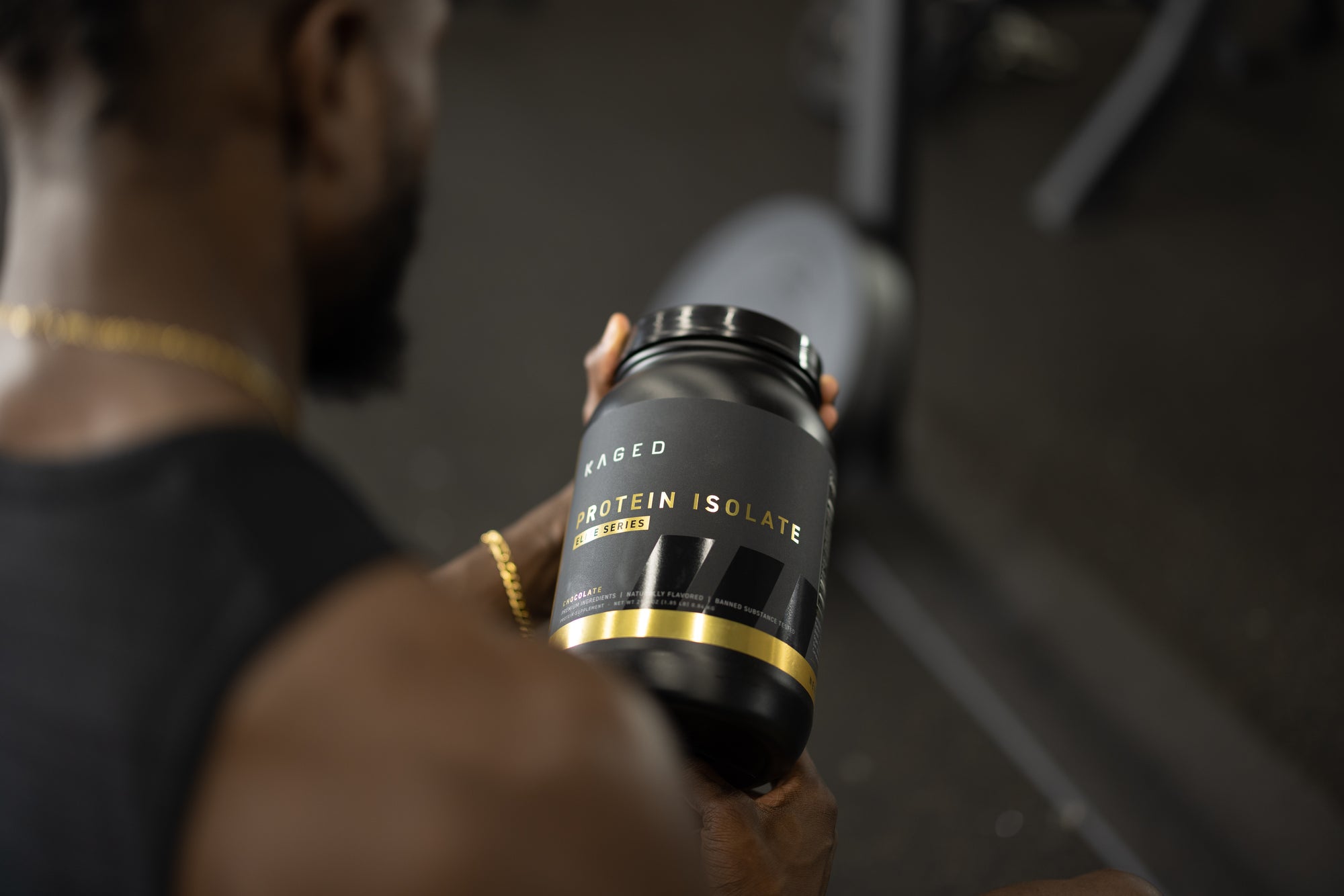 Angled view of KAGED Protein Isolate tub showcasing black-and-gold label reading Protein Isolate with blurred gym equipment background