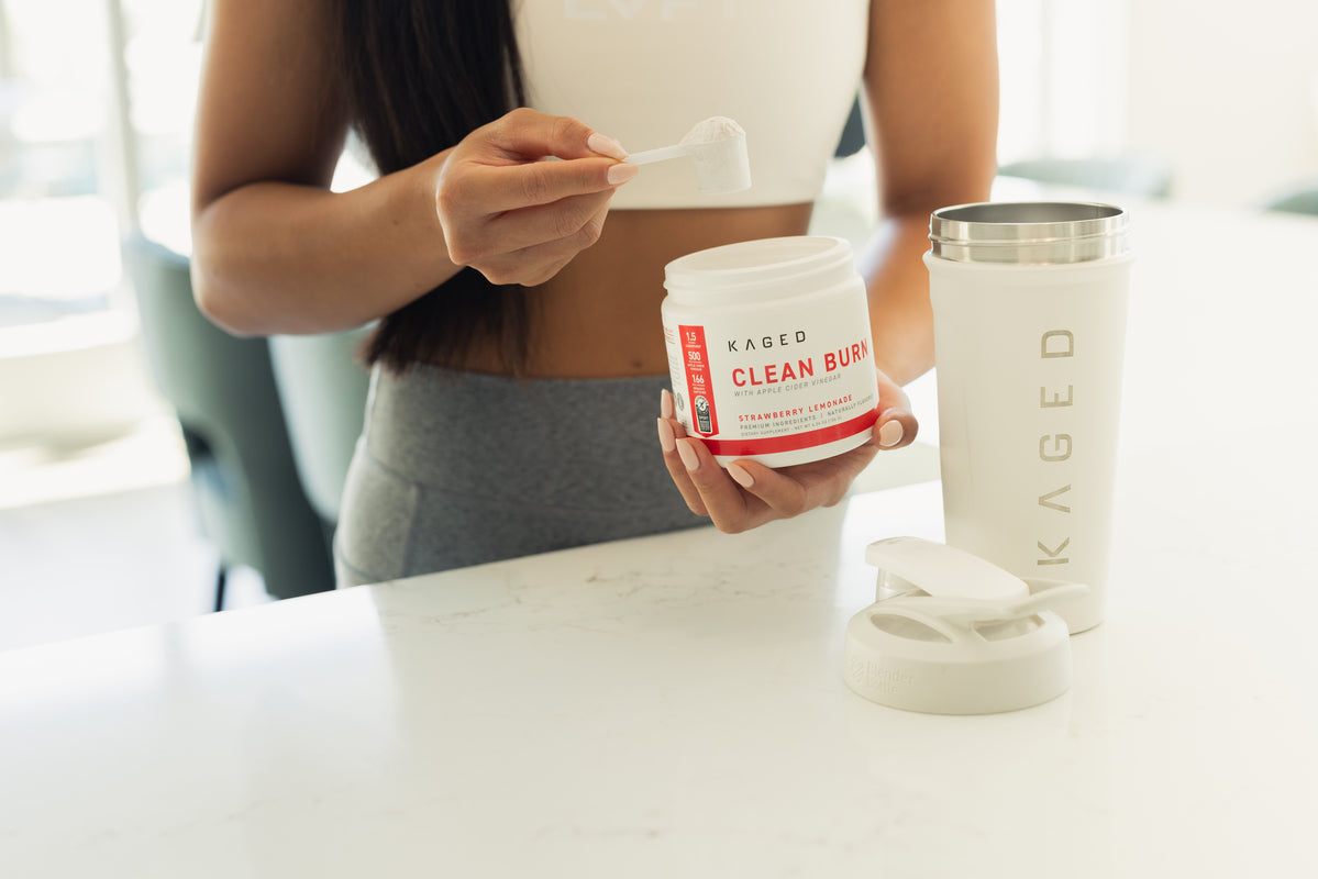 Woman scooping KAGED Clean Burn supplement powder in strawberry lemonade flavor into a scoop beside a white KAGED shaker on a kitchen counter