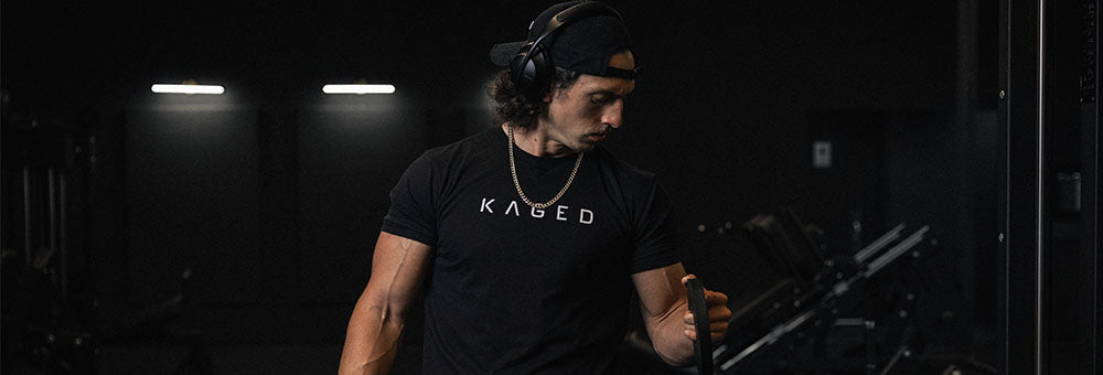 Man wearing KAGED t-shirt and headphones adjusting gym equipment in a dimly lit weight room